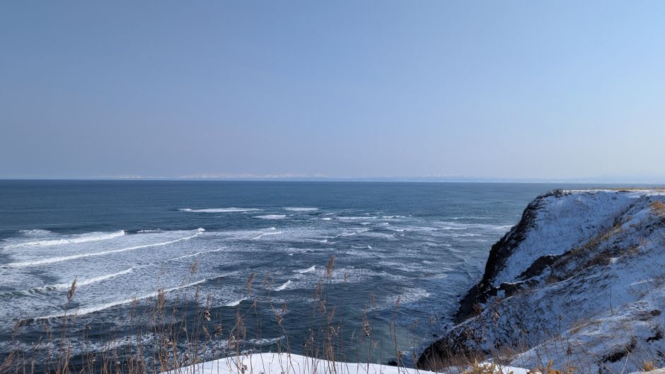 The white lighthouse at Cape Chikyū in Muroran standing above sheer green cliffs that plunge into a deep blue sea under a windy sky