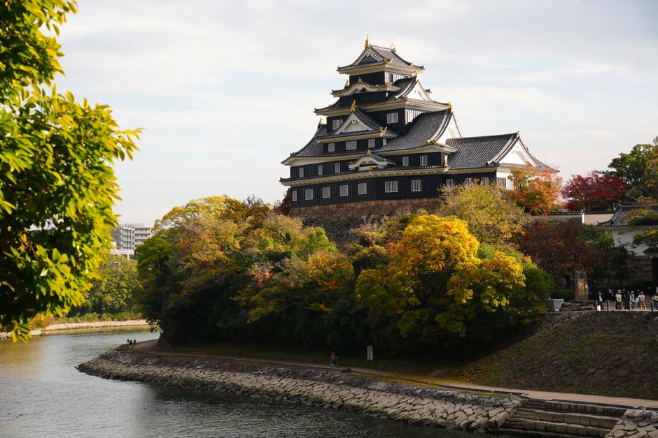 Wide lawns and a still pond in Kōraku-en garden, clipped pines and a small wooden pavilion, the black Okayama Castle visible above the trees in golden evening light