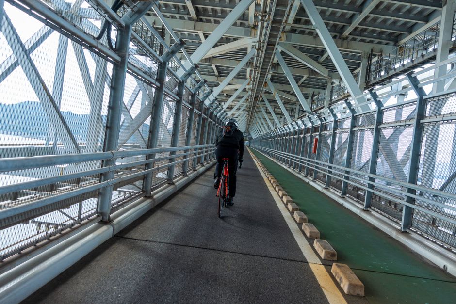 A cyclist on the Shimanami Kaidō route near Onomichi, the dedicated bike lane running beside the calm Inland Sea toward a great white suspension bridge spanning to a green island