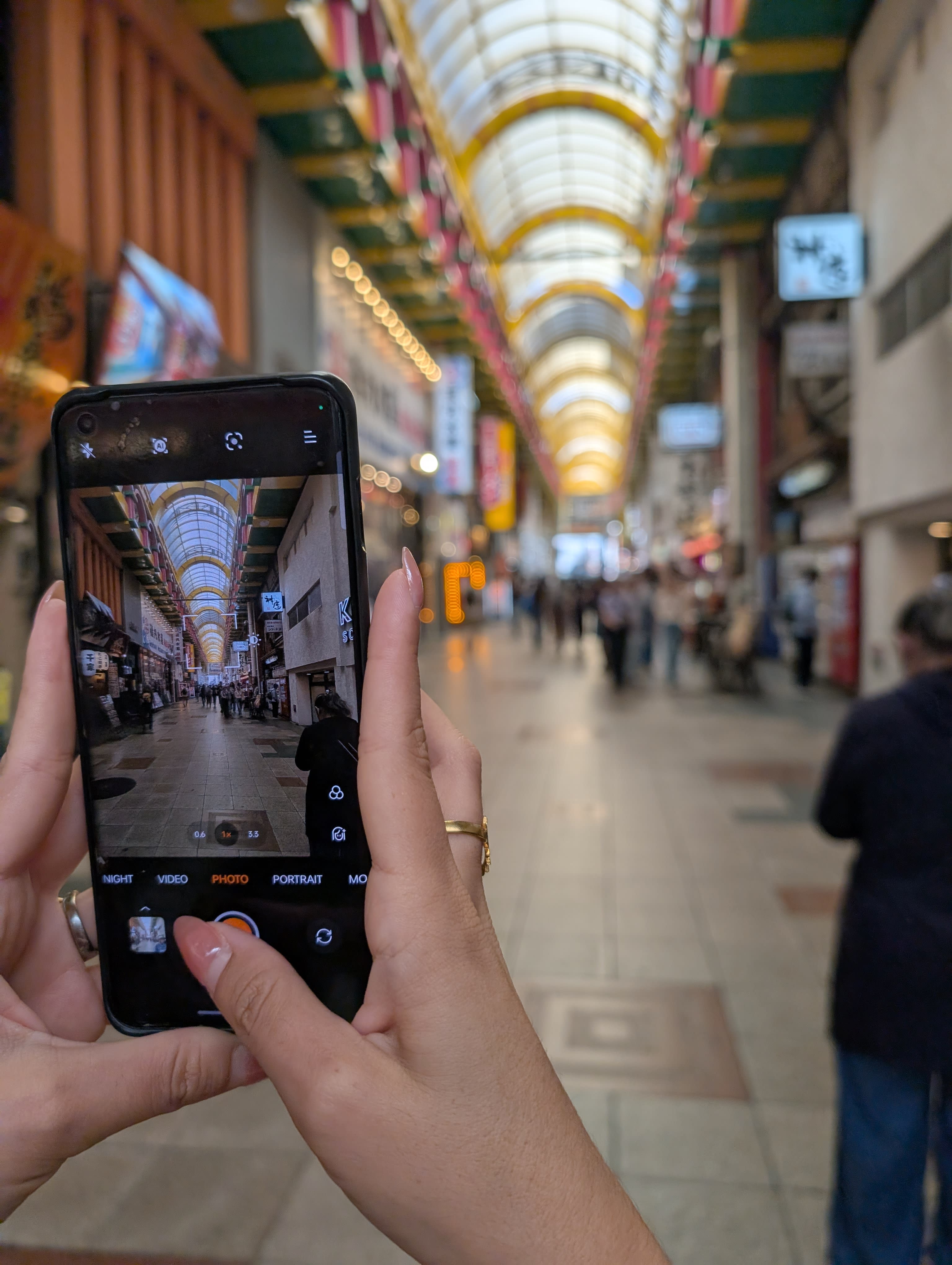 Lia photographing the endless Shinsaibashi shopping arcade — the arched glass ceiling, the lights, the crowd stretching into the distance captured on her phone screen