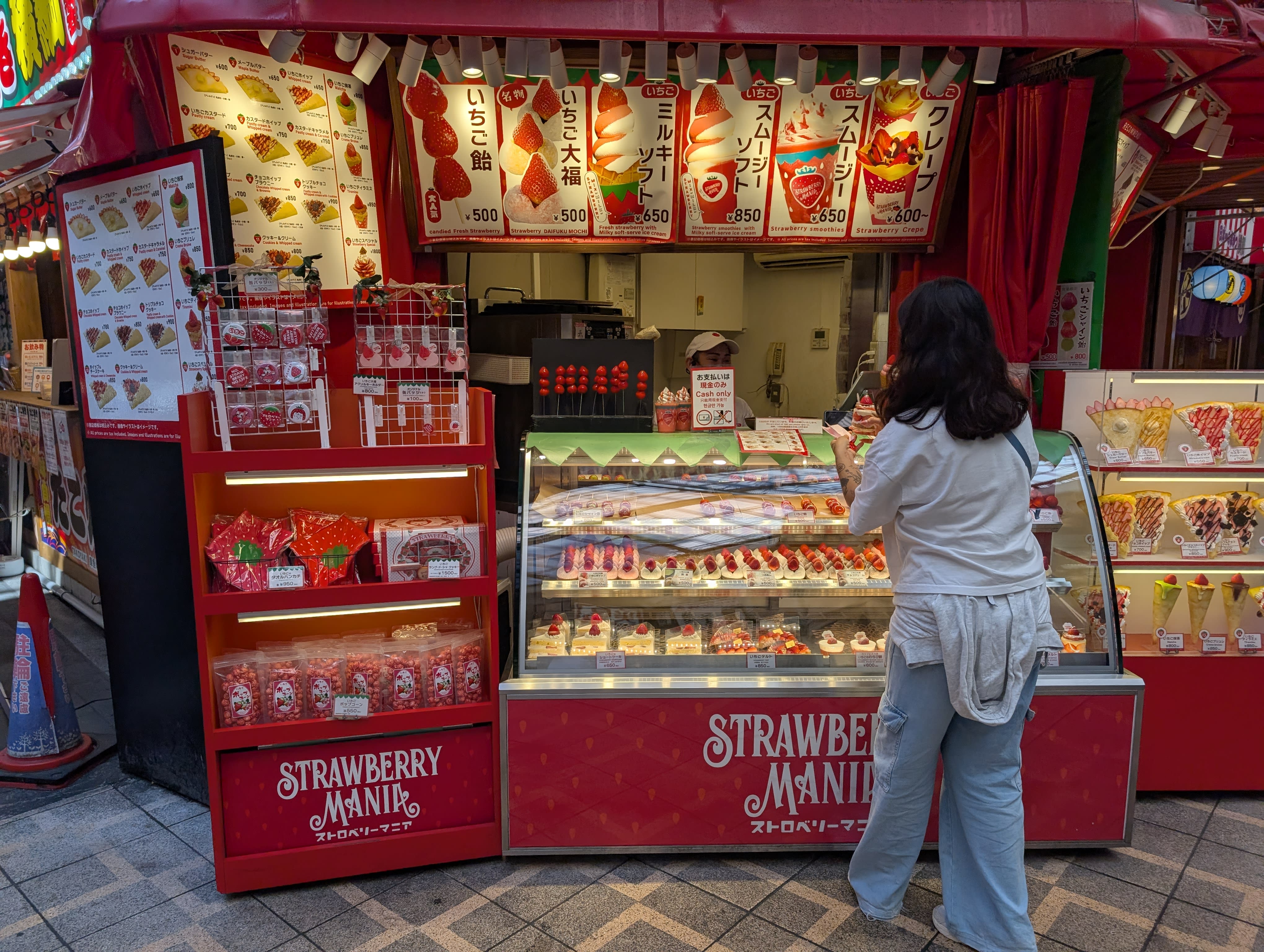 Lia at Strawberry Mania in the Shinsaibashi arcade — the red-and-gold stall packed with strawberry desserts, her back to the camera, choosing carefully