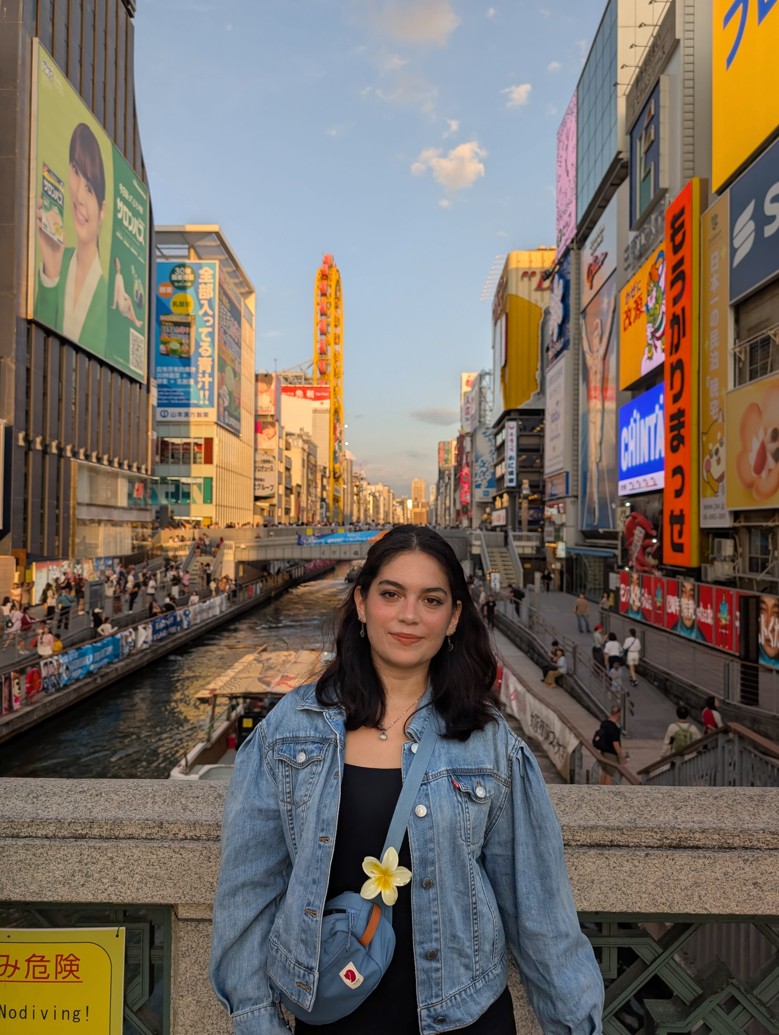 Lia on the Dotonbori bridge at golden hour — the canal stretching behind her, neon signs on both banks, the city shifting from afternoon to evening