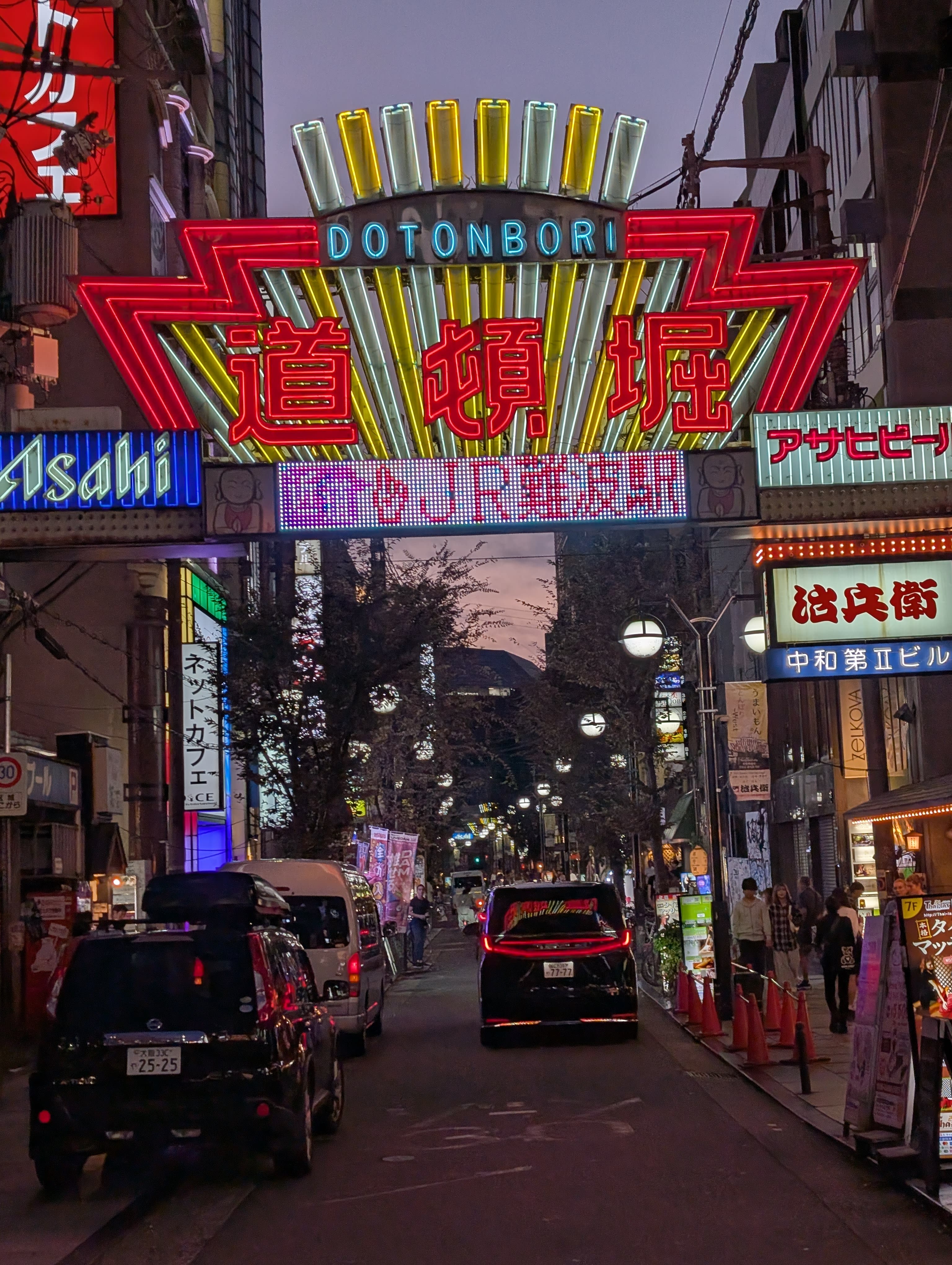 The Dotonbori gate at dusk — neon kanji glowing red and yellow, Asahi signs, cars passing beneath into the electric heart of Osaka's night