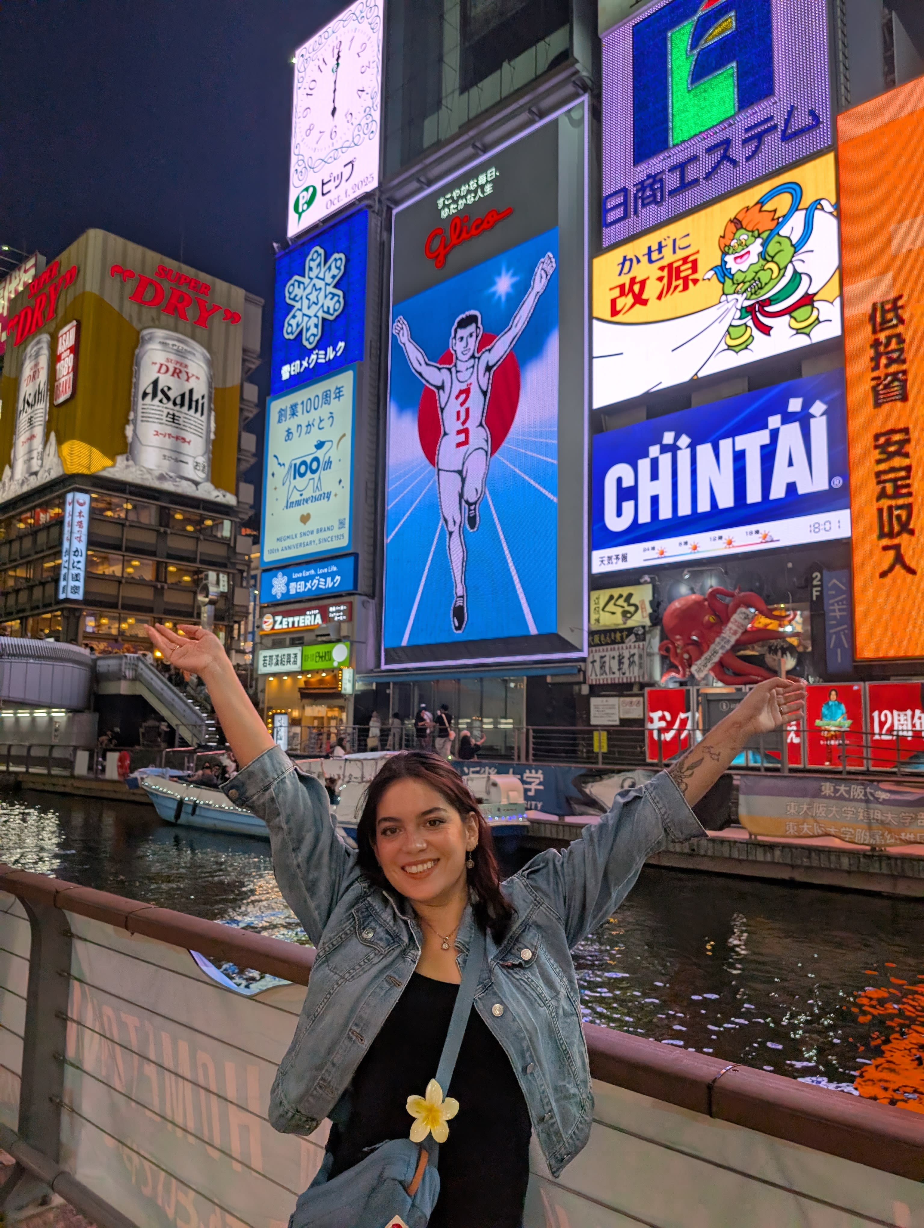 Lia doing the Glico Running Man pose on the Dotonbori bridge at night — arms raised, grinning, the iconic neon billboard perfectly framed behind her