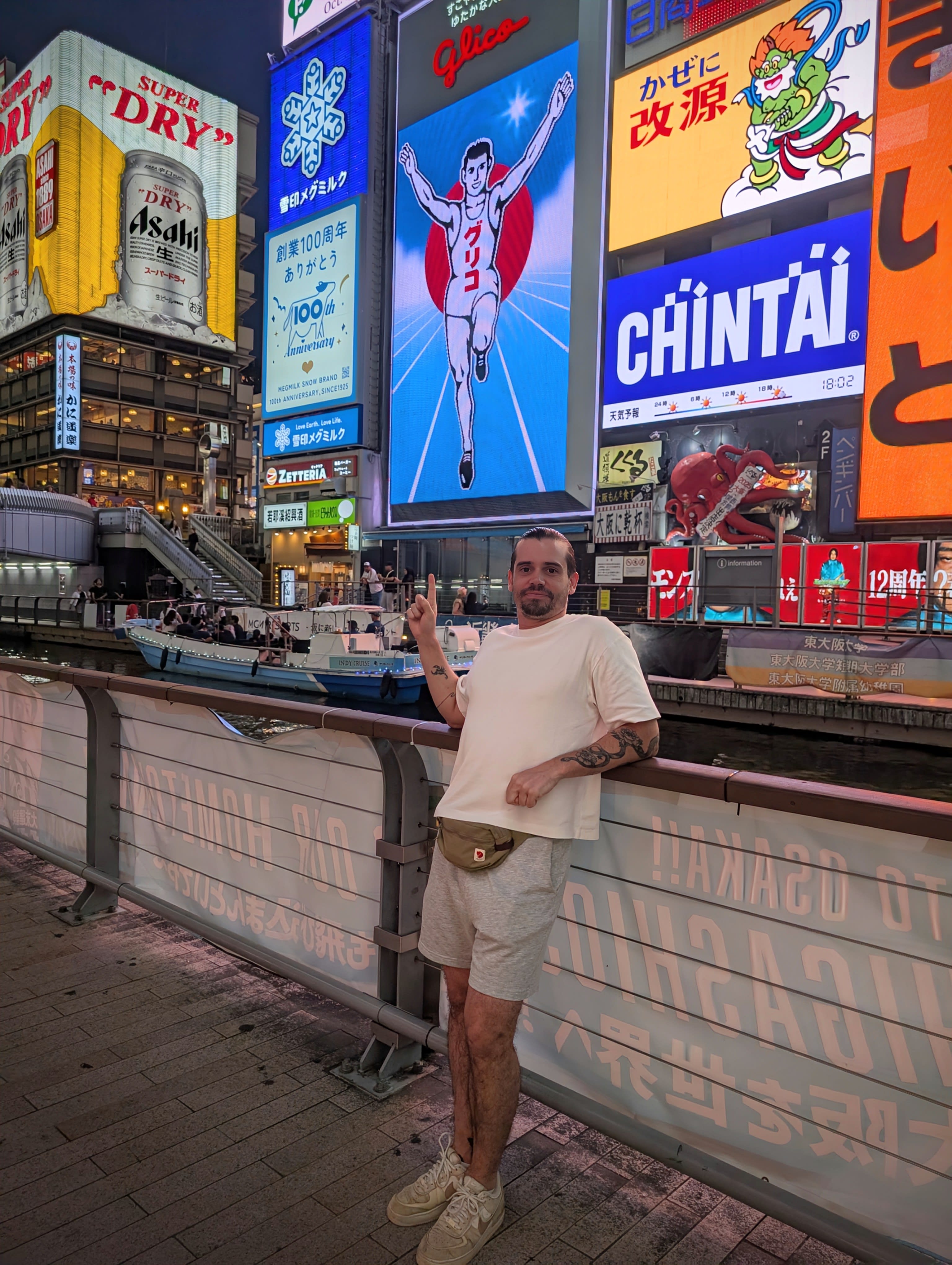 Pierre in front of the Glico Running Man sign at night — peace sign, the neon reflecting on the canal, a boat passing behind him