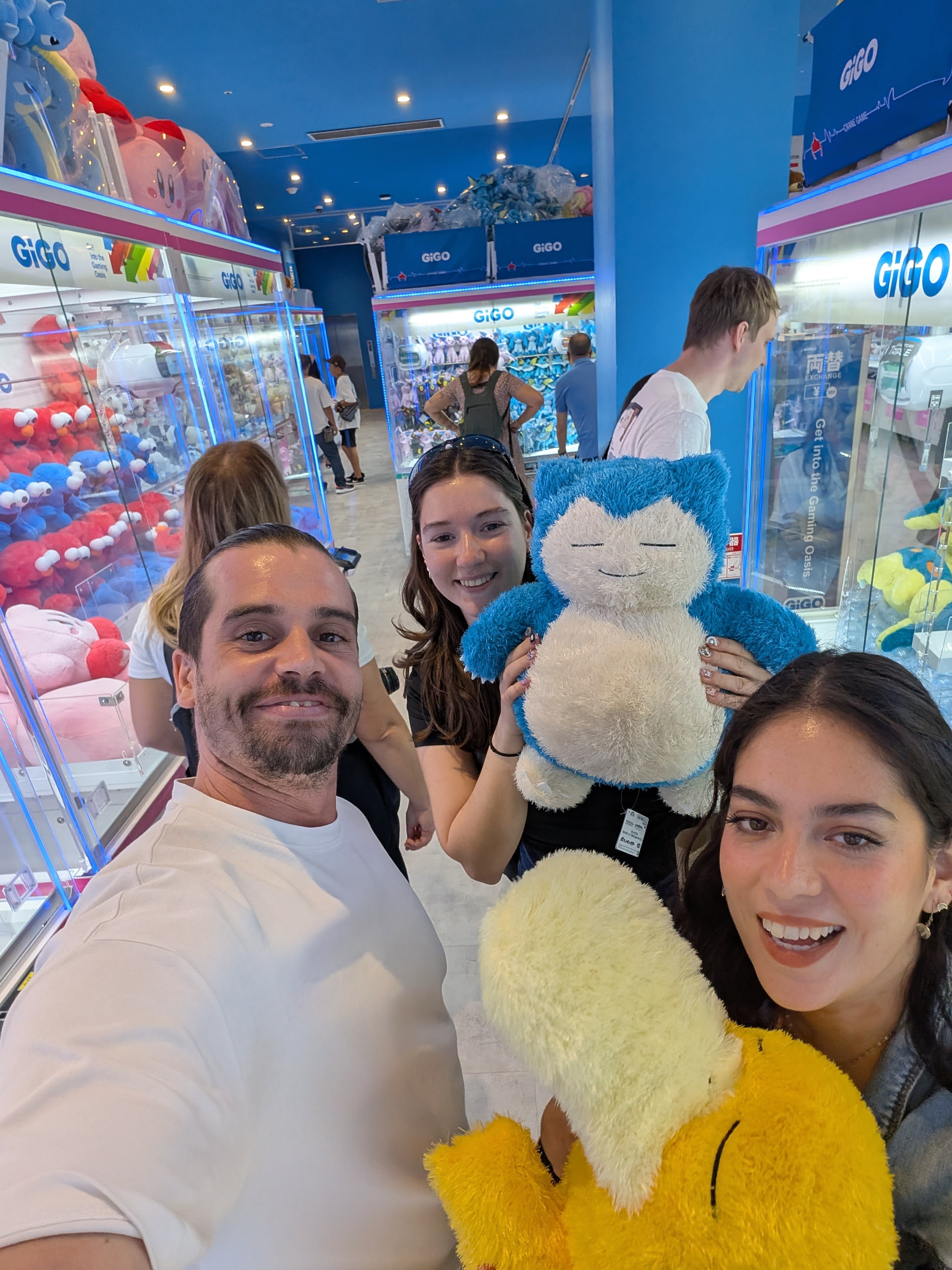 Pierre, a fellow traveler, and Lia at GiGO — holding giant Pokémon plushies won from the crane machines, the blue-lit arcade glowing behind them