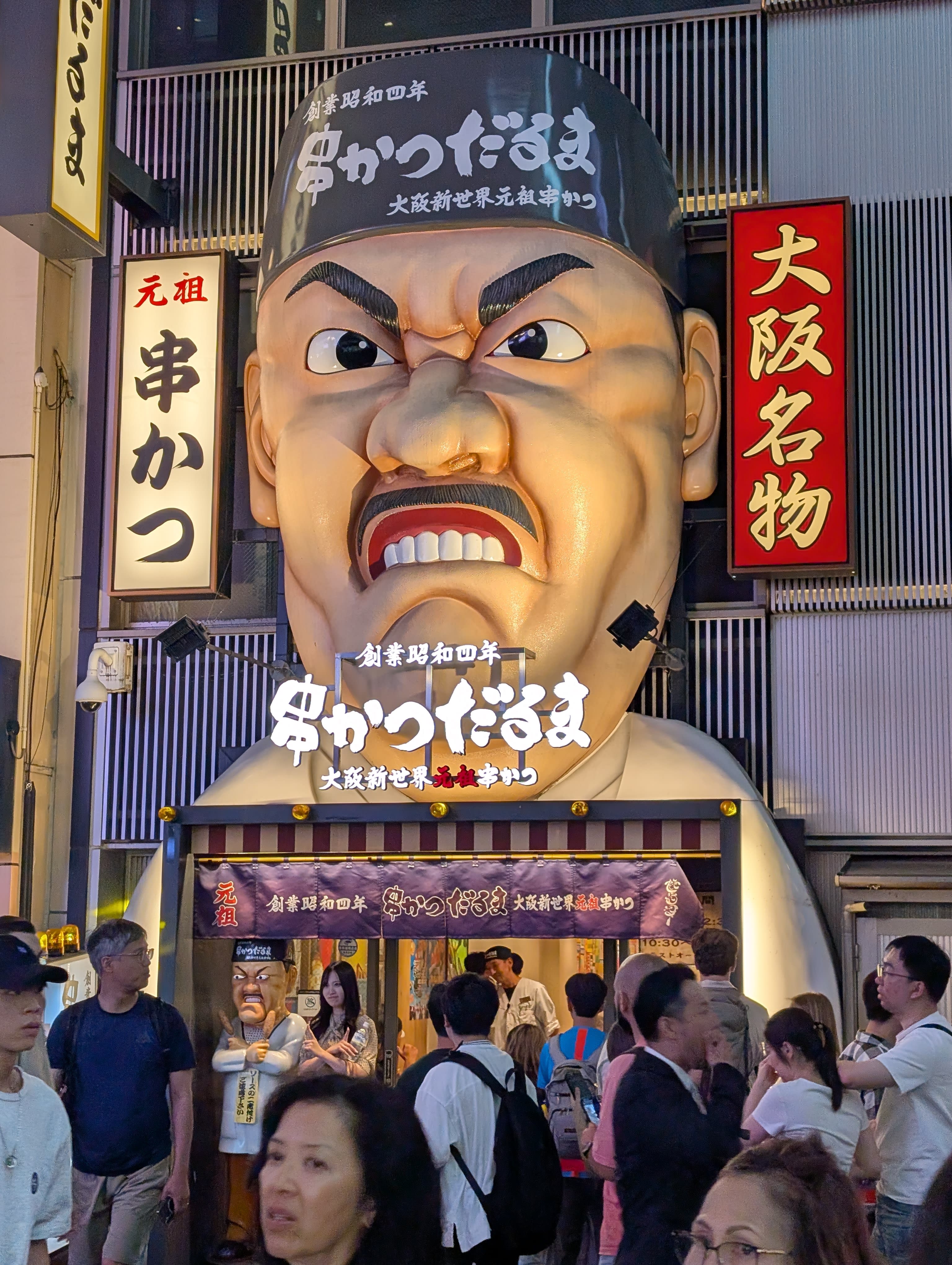 The iconic Kushikatsu Daruma restaurant — the giant angry-faced statue looming over the entrance, the crowd flowing past, kanji signs stacked above