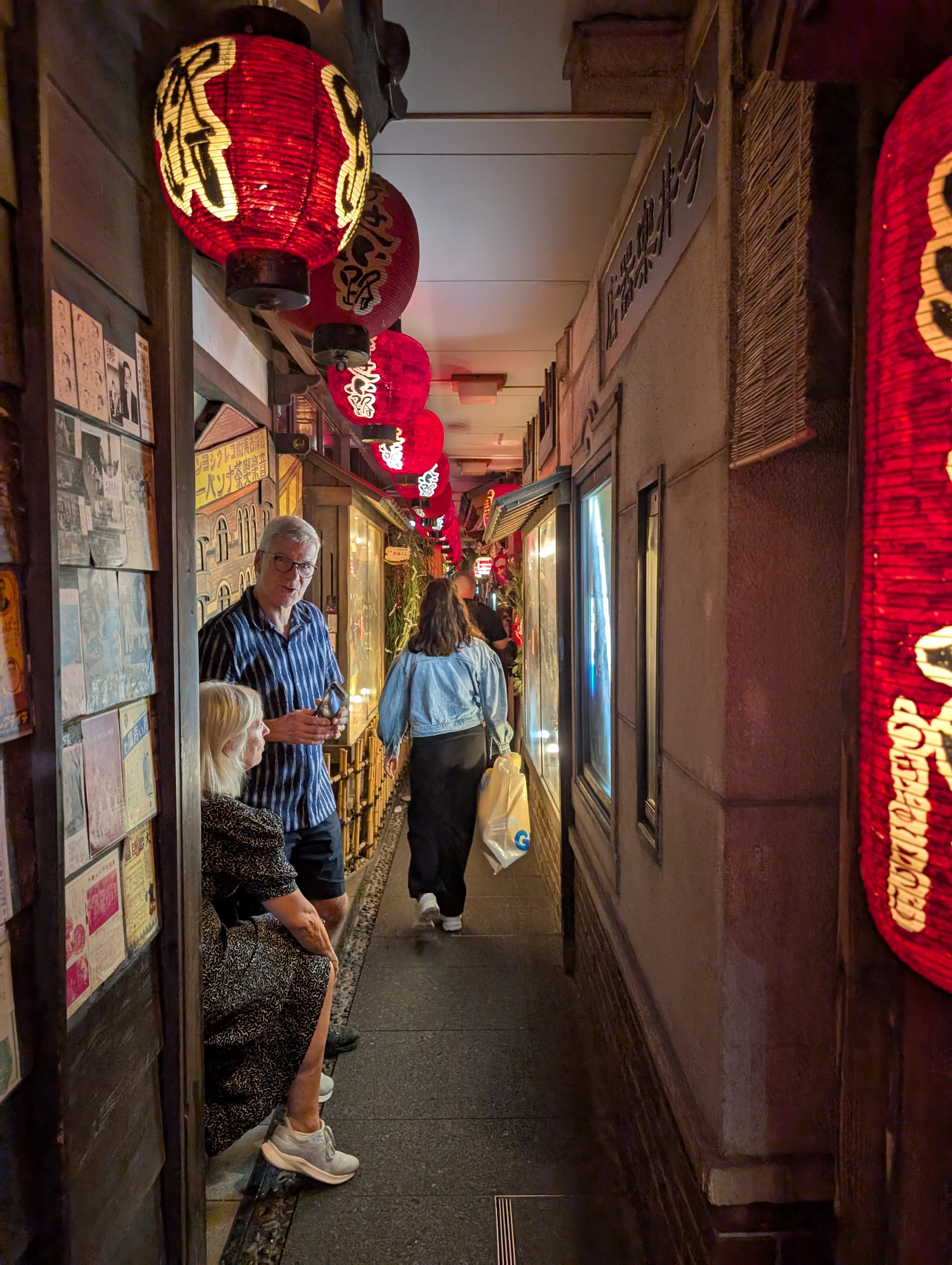 A narrow alley behind Dotonbori — red paper lanterns hanging overhead, a couple walking through, the warm glow of tiny restaurants on both sides
