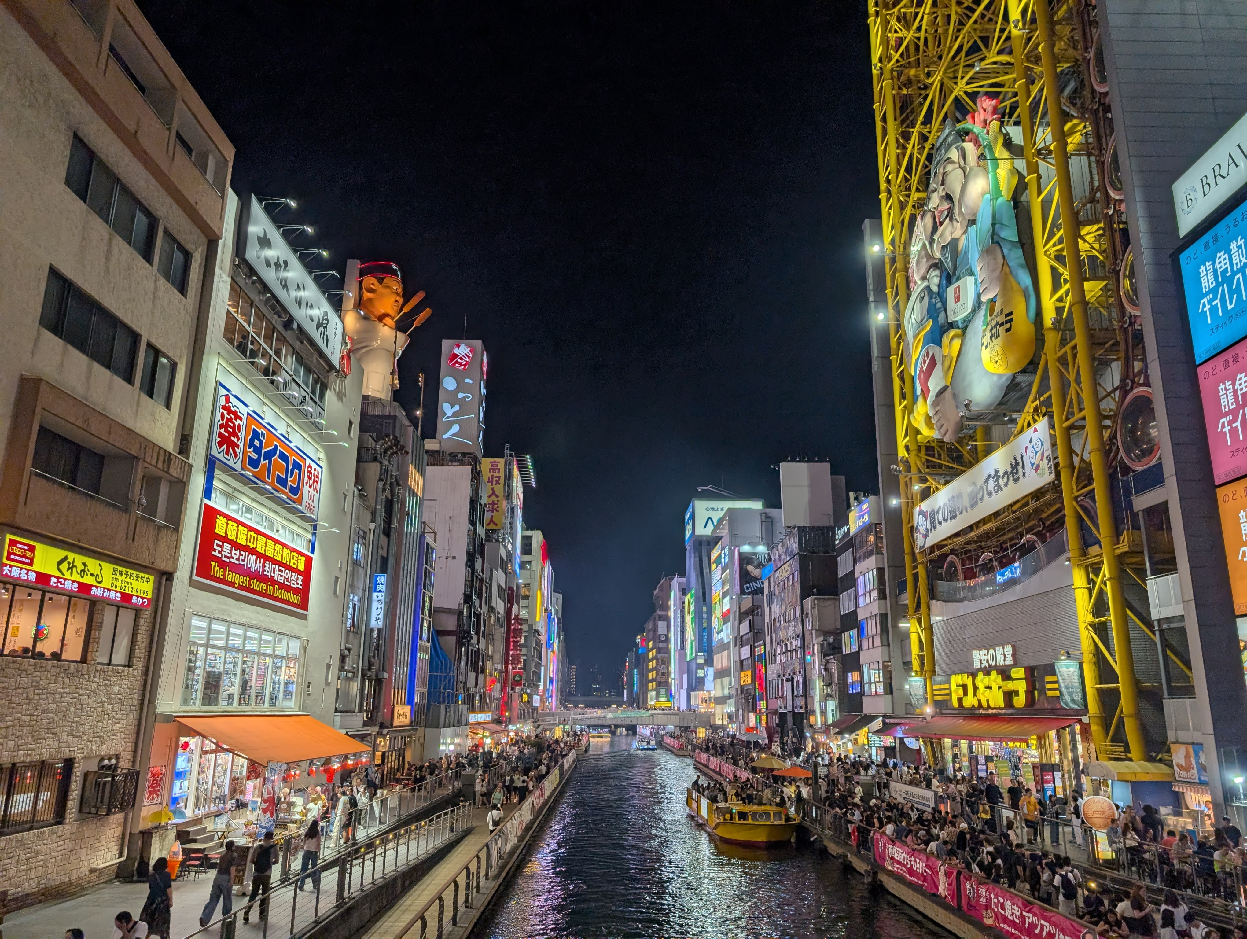 The Dotonbori canal at night from the bridge — neon blazing on both banks, a tour boat gliding through, the entire scene reflected in the dark water below