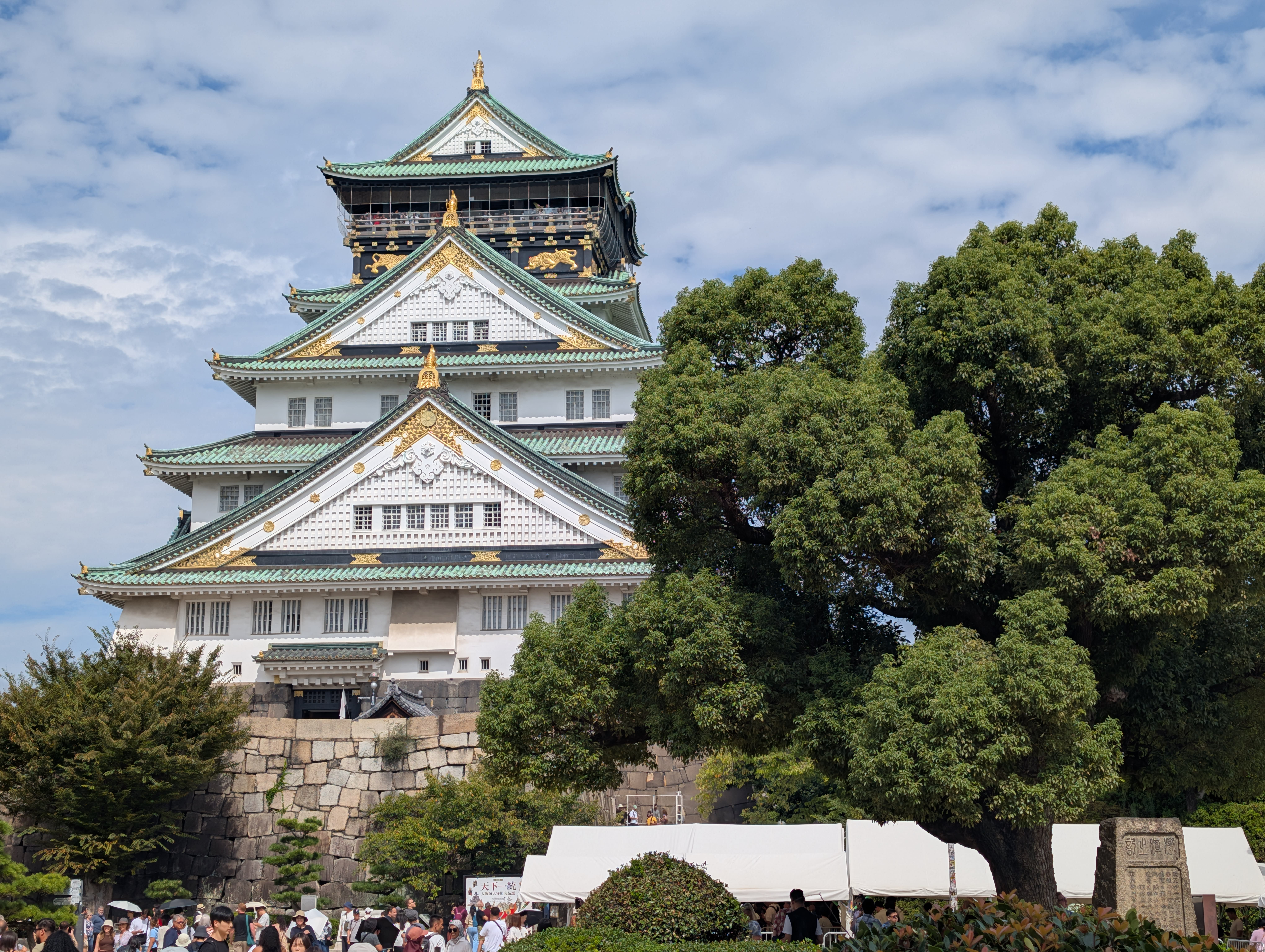 Osaka Castle in full — the white walls, the green-and-gold rooftops, the ancient trees flanking it, the stone walls rising from the moat below