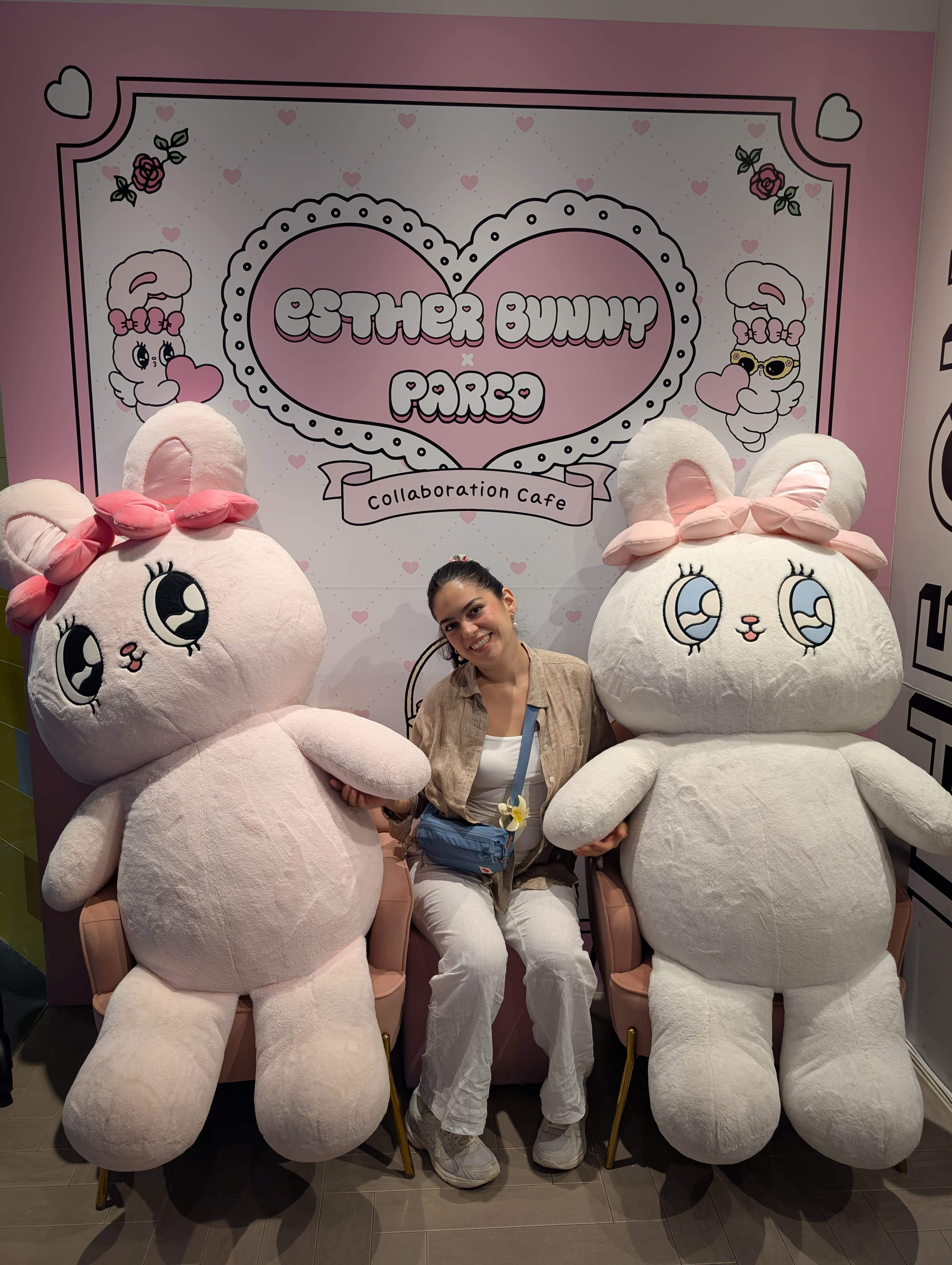 Lia at the Esther Bunny Café at Parco — seated between two giant white stuffed bunnies, pink walls behind her, looking entirely content