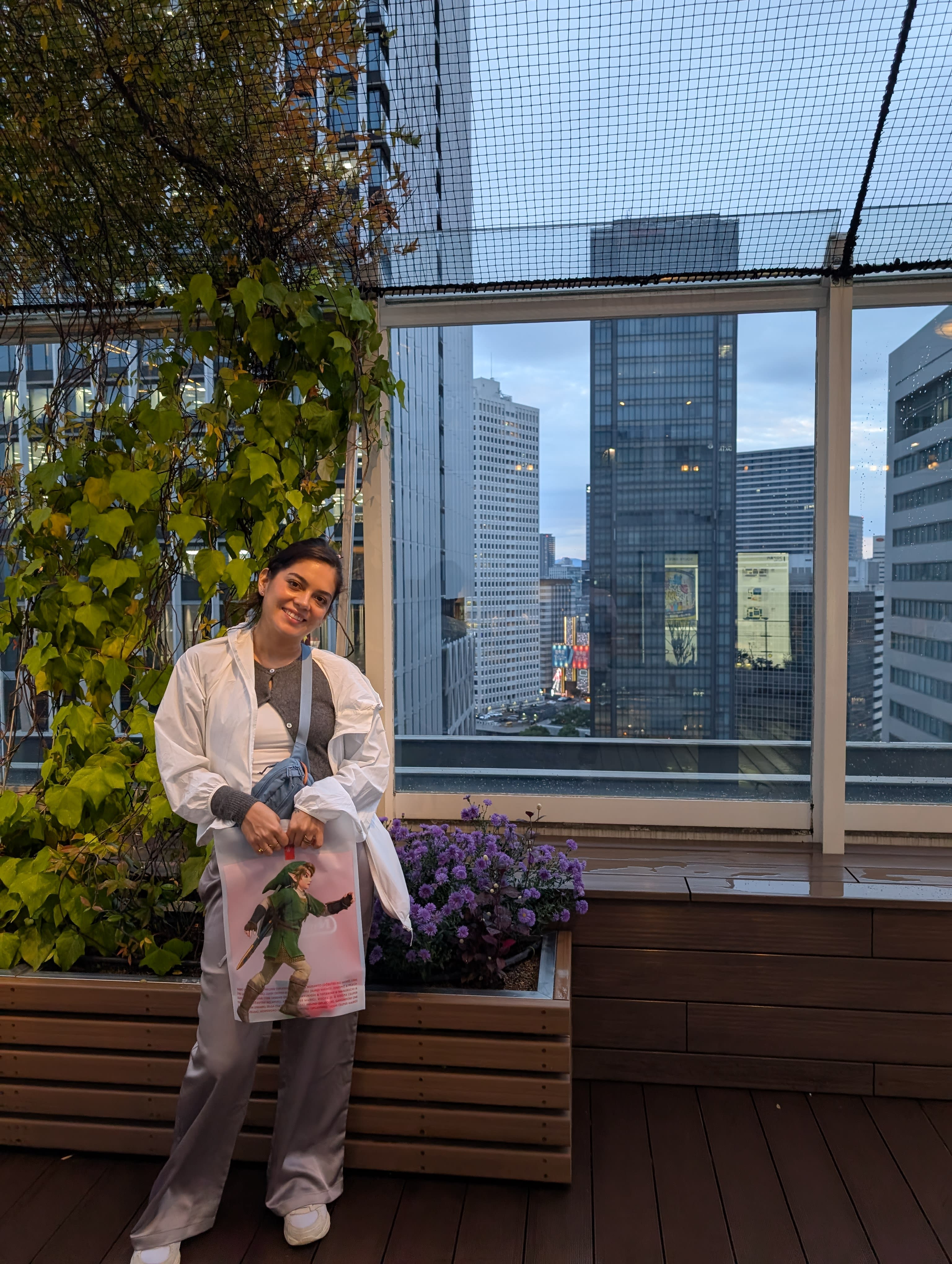 Lia on a rooftop terrace in Umeda — skyscrapers behind her, purple flowers beside her, the city stretching in every direction, her expression somewhere between home and wonder