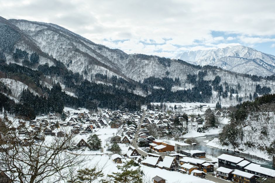 A gasshō-zukuri farmhouse lit warmly from within at dusk, glowing across the darkening rice paddies of Shirakawa-gō