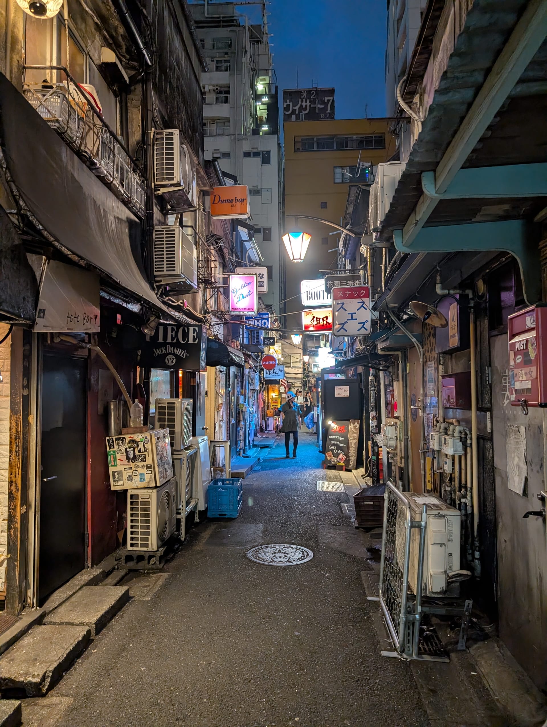 A narrow Golden Gai alley at night — tiny bars on both sides, neon signs, air conditioning units, and a lone figure in the blue-lit distance