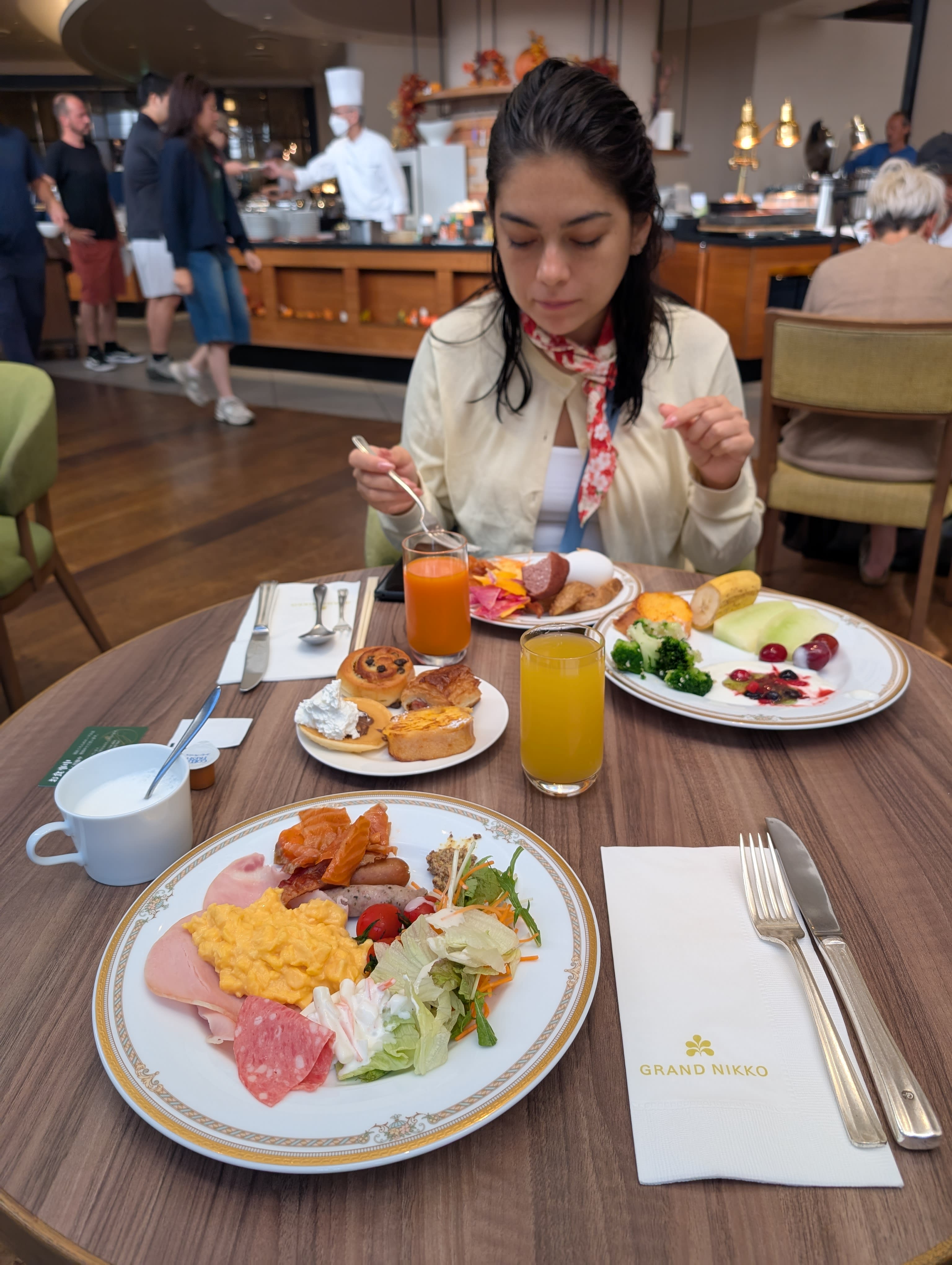 Lia at the Grand Nikko breakfast buffet — two plates loaded, orange juice, pastries, the open kitchen and morning light behind her