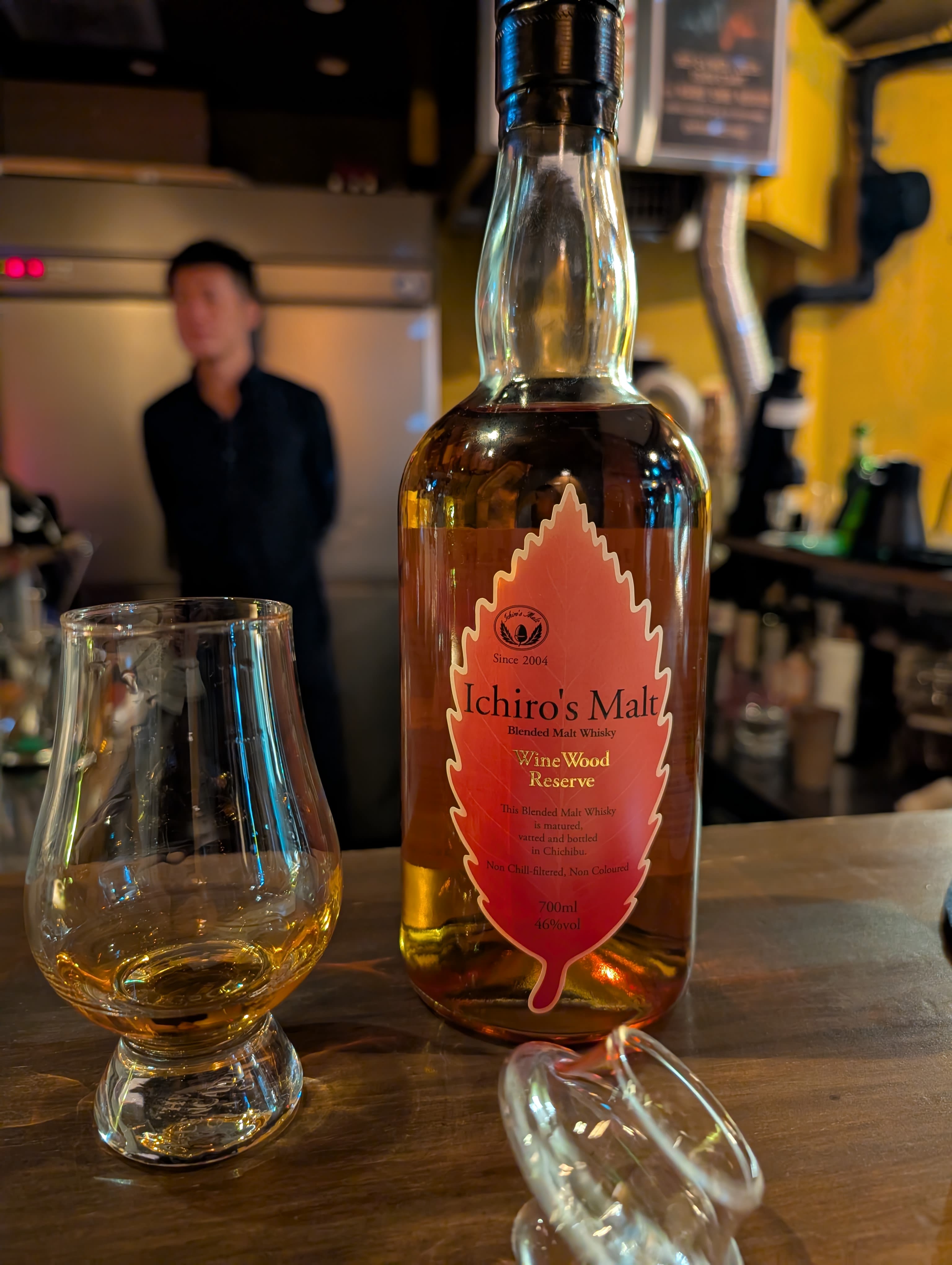 A bottle of Ichiro's Malt Wine Wood Reserve whisky with a Glencairn glass at a Tokyo whisky bar — amber liquid, soft lighting, the bartender a blur behind
