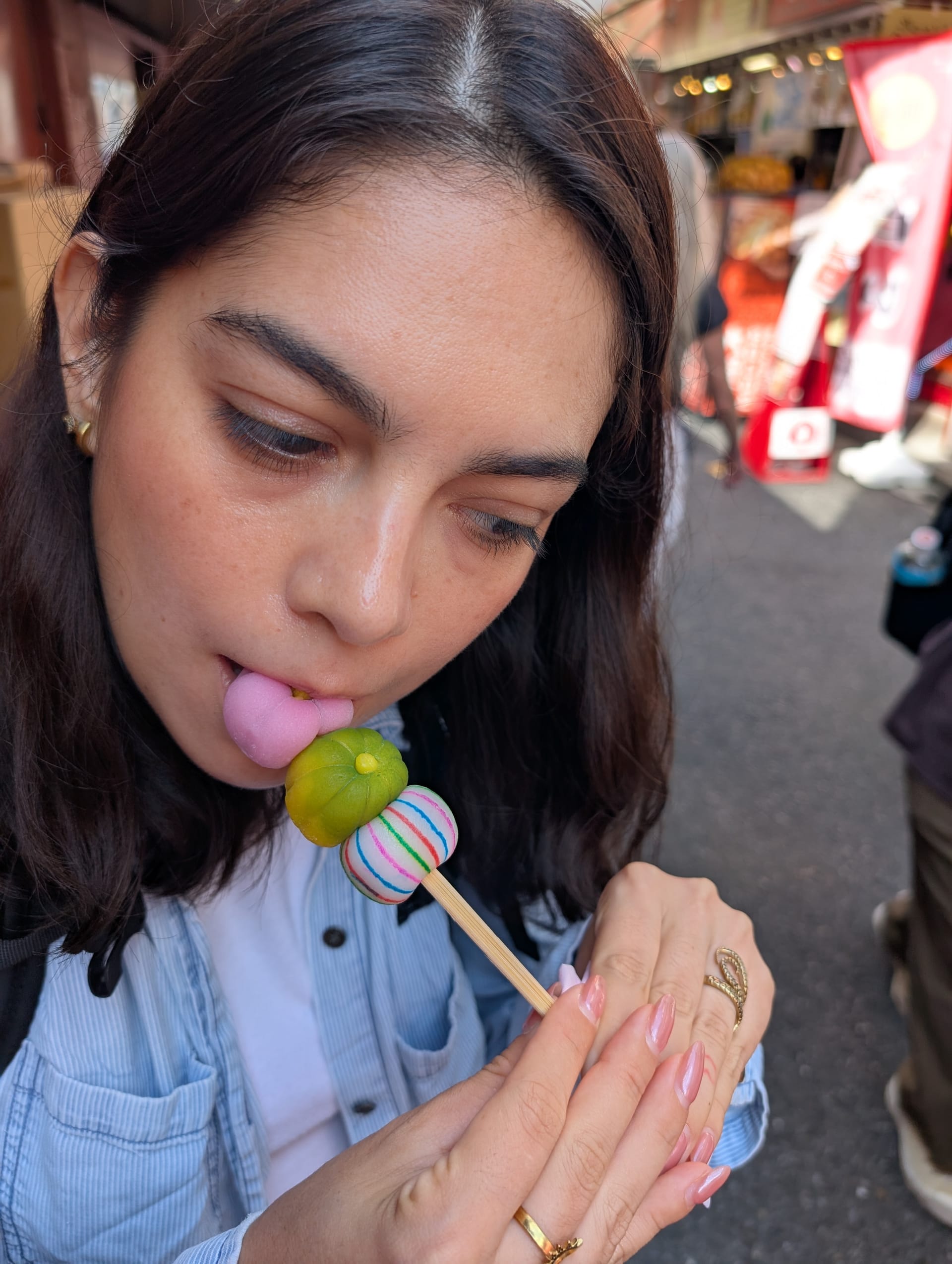 Lia eating dango on a skewer in Asakusa — pink and green mochi balls, the market stalls blurred behind her