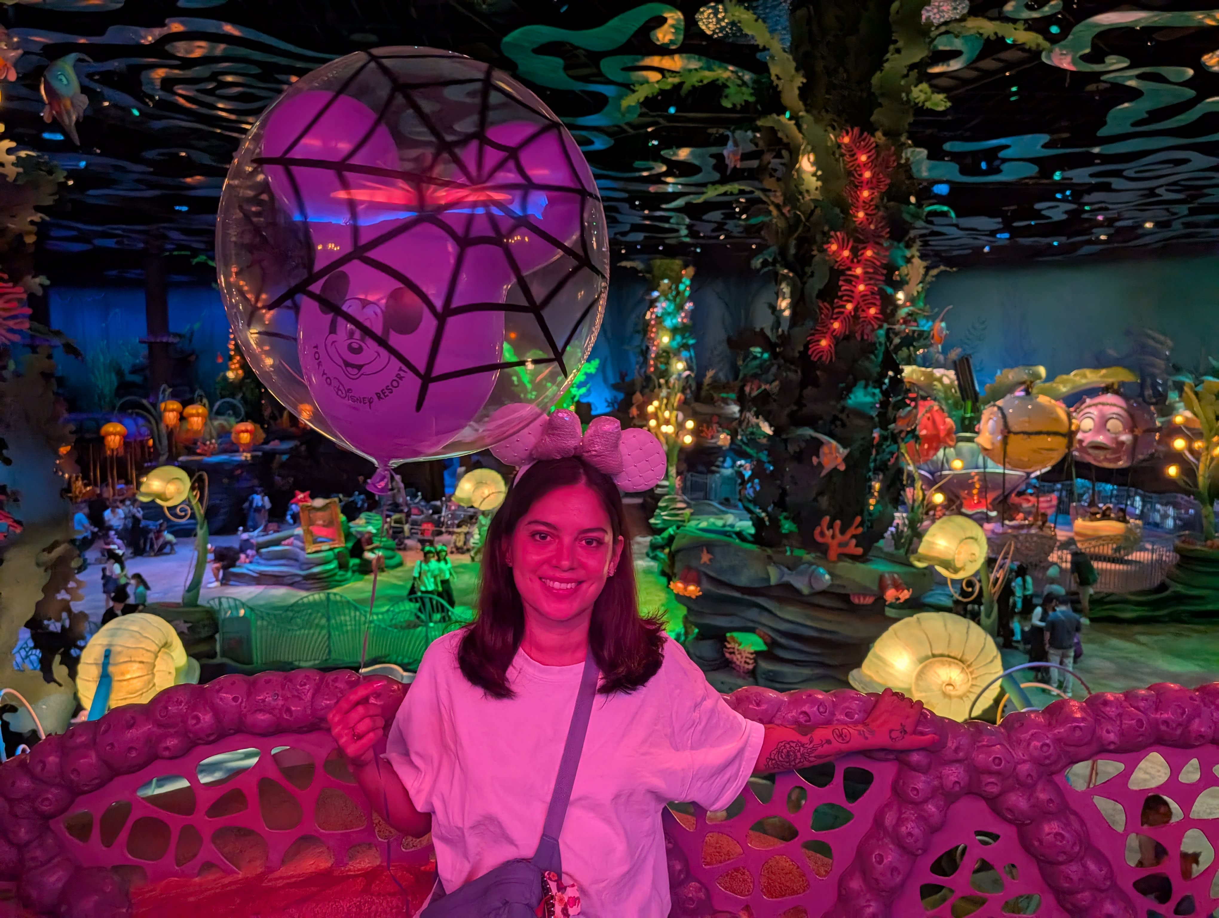 Lia inside DisneySea's Mermaid Lagoon — purple and green light, an underwater fantasy world of shells and coral glowing around her, a Halloween balloon overhead