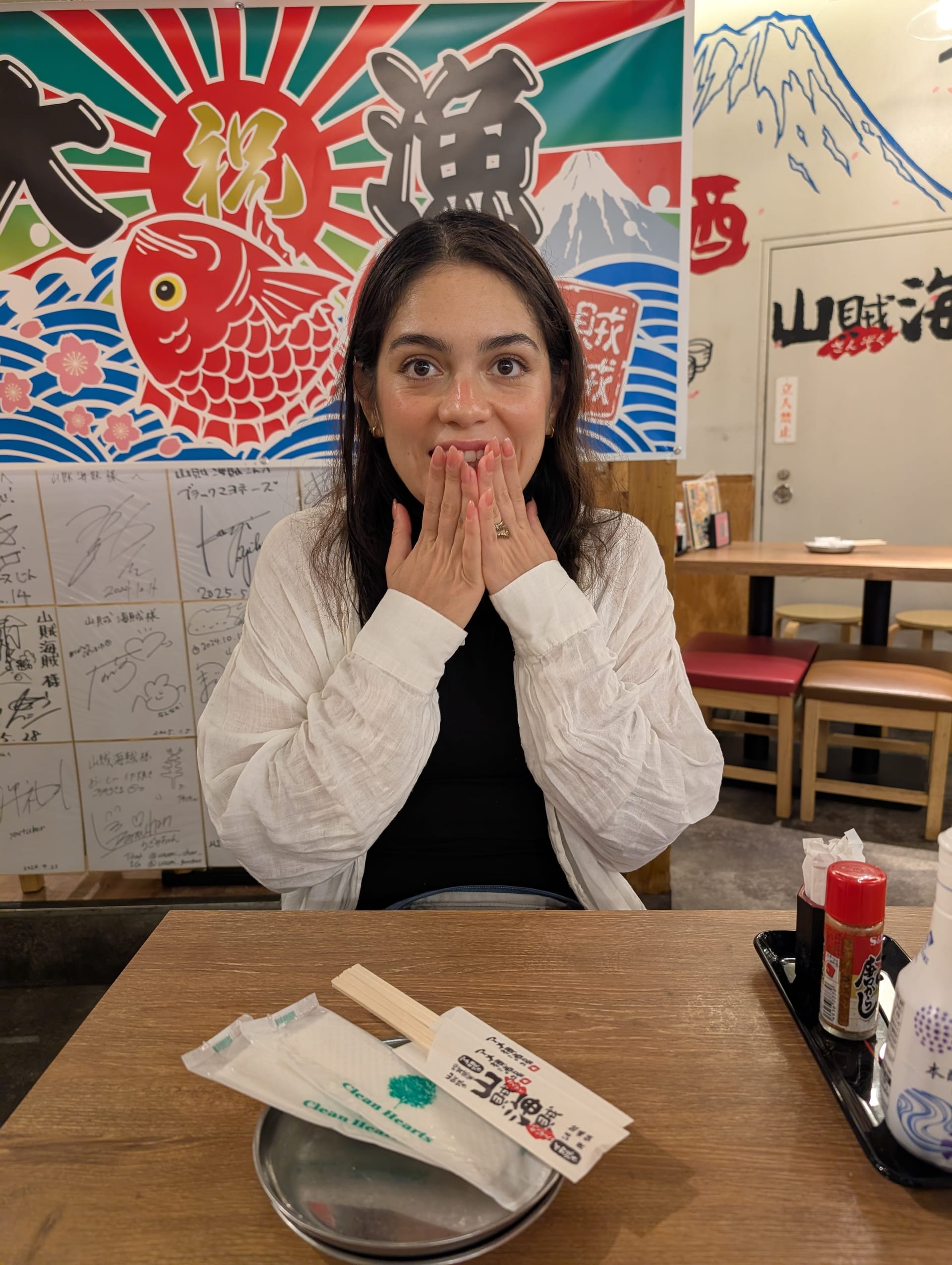 Lia in our first Tokyo izakaya — hands over her mouth in that expression she makes when she is simultaneously overwhelmed and delighted, a painted fish mural behind her