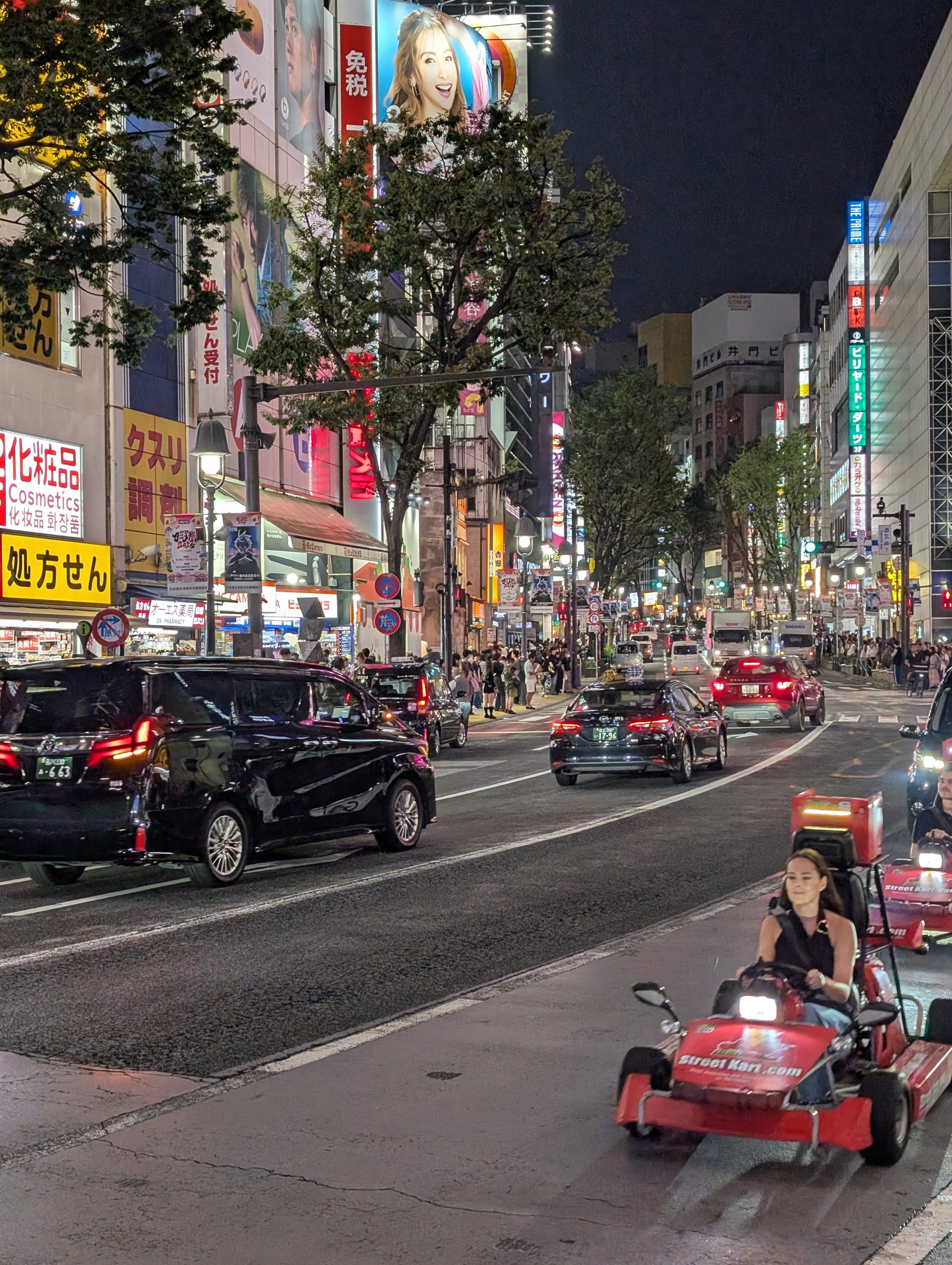 Lia driving a go-kart through Shibuya at night — red kart, neon signs, traffic, the city blazing around her