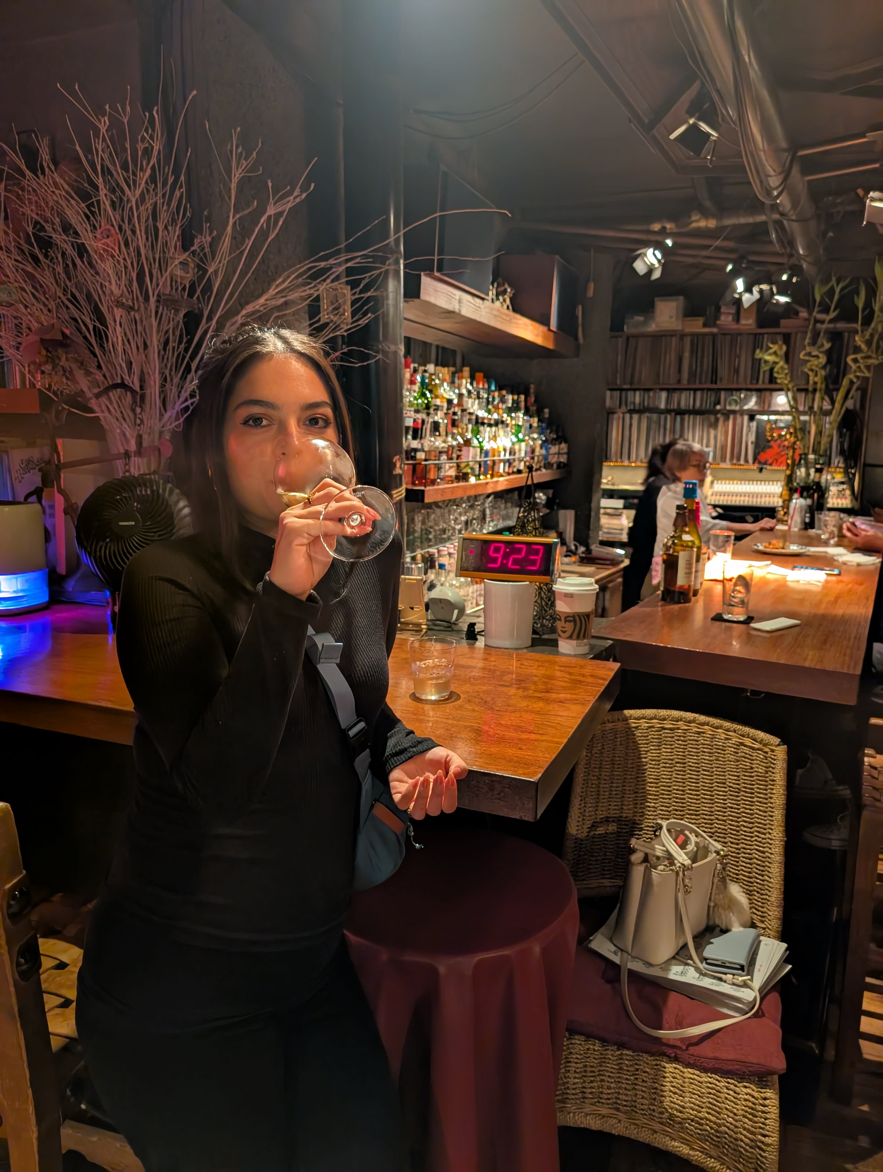 Lia sipping wine at a Roppongi bar — warm lighting, vinyl records stacked on shelves, a red digital clock reading 9:29