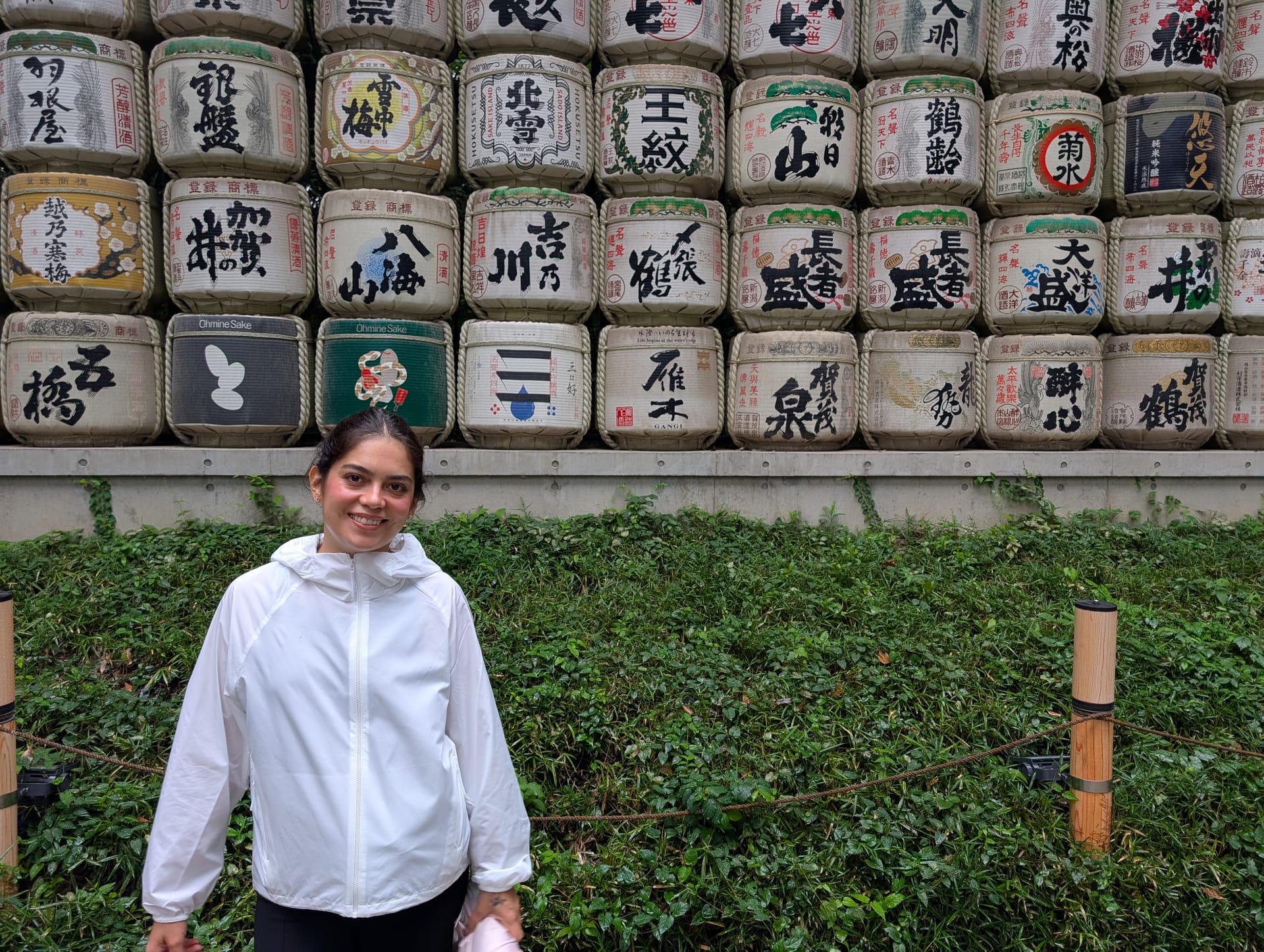 Lia standing in front of the sake barrel display at Meiji Jingu — rows of ceremonial barrels with kanji labels rising behind her