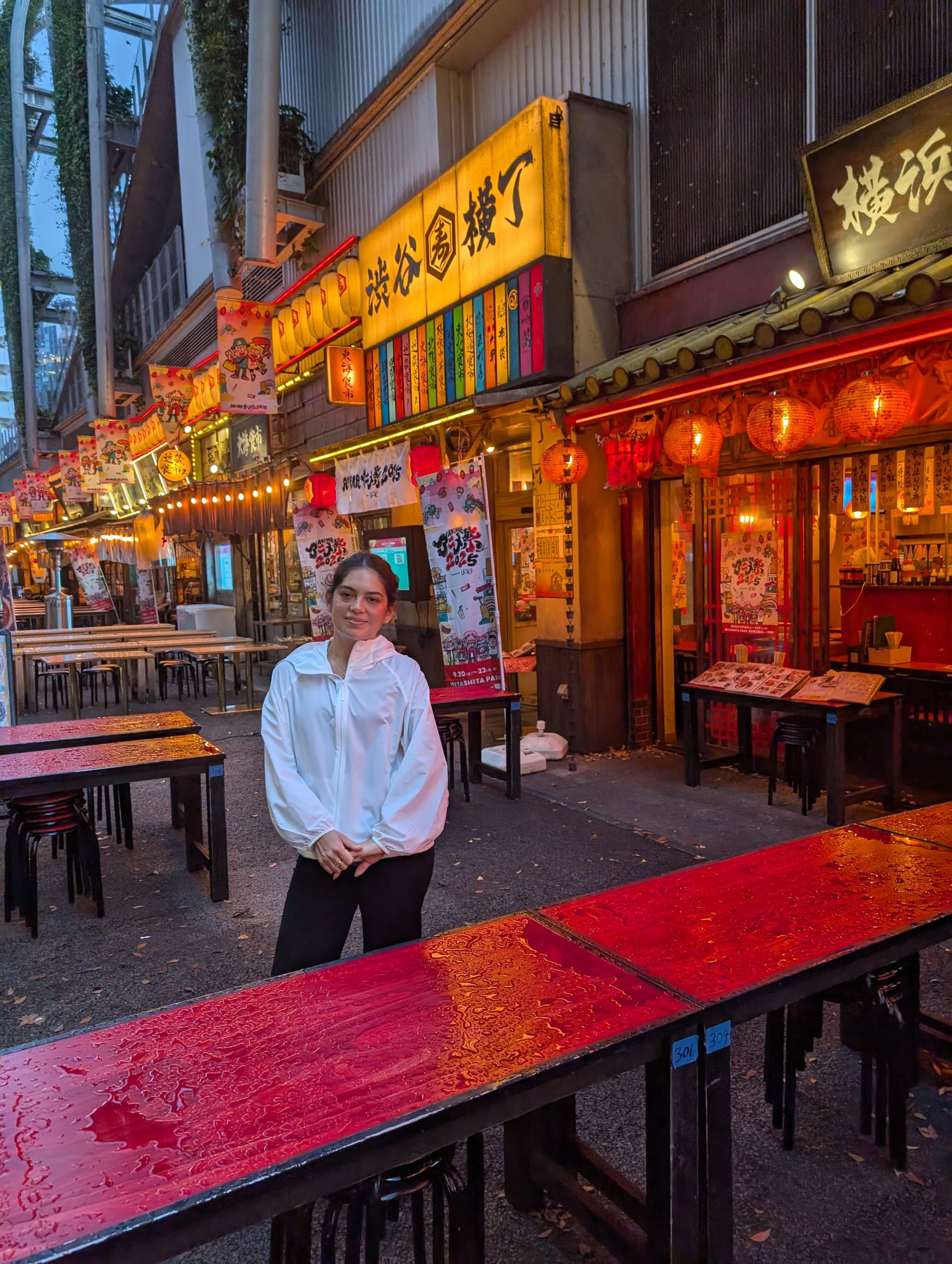 Lia standing at the entrance of Shibuya Yokocho — red lanterns, wet wooden tables, and the warm glow of food stalls stretching behind her