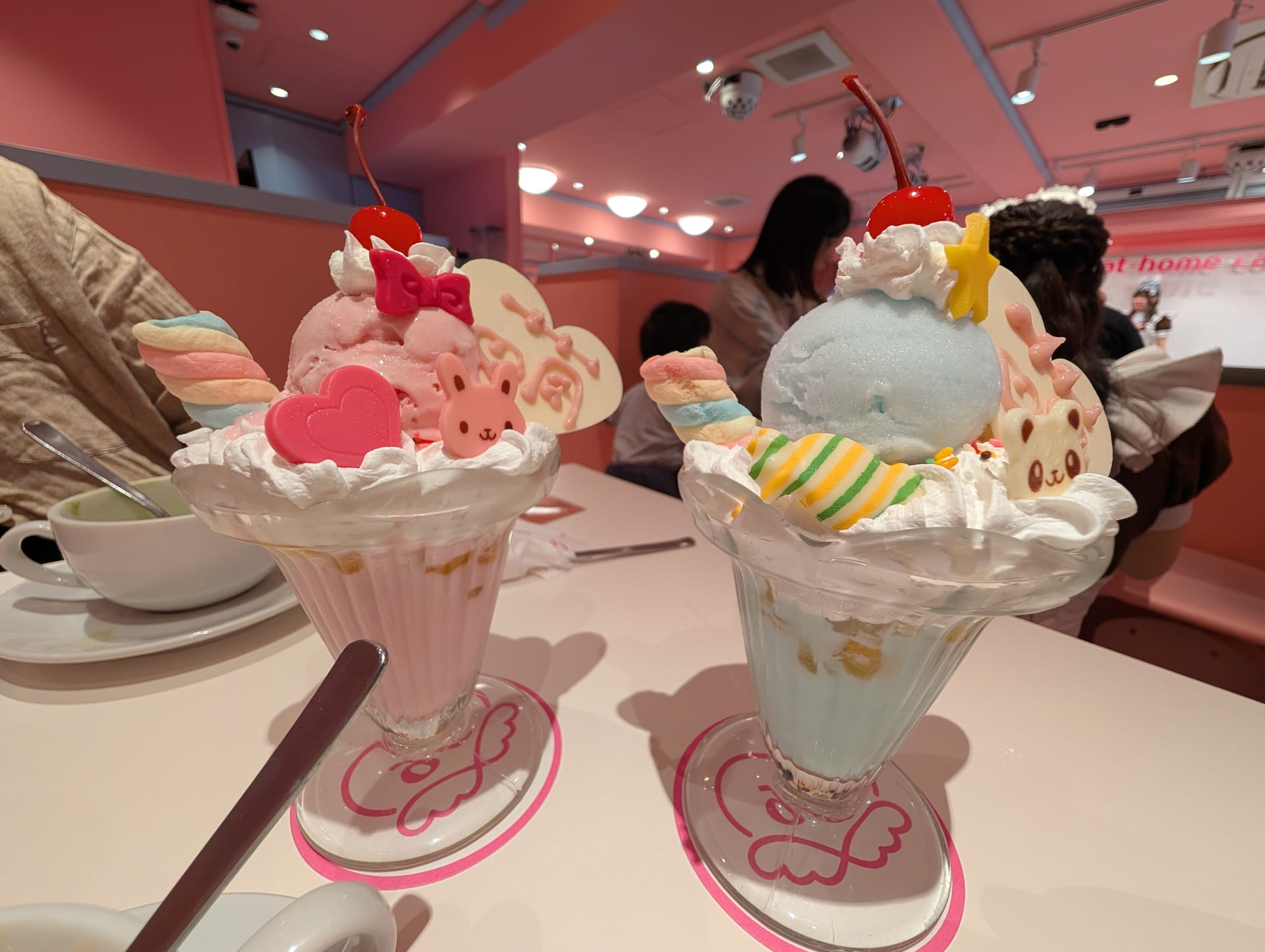 Two kawaii parfaits at a maid cafe — towers of pastel ice cream, character cookies, hearts, and whipped cream in pink glasses