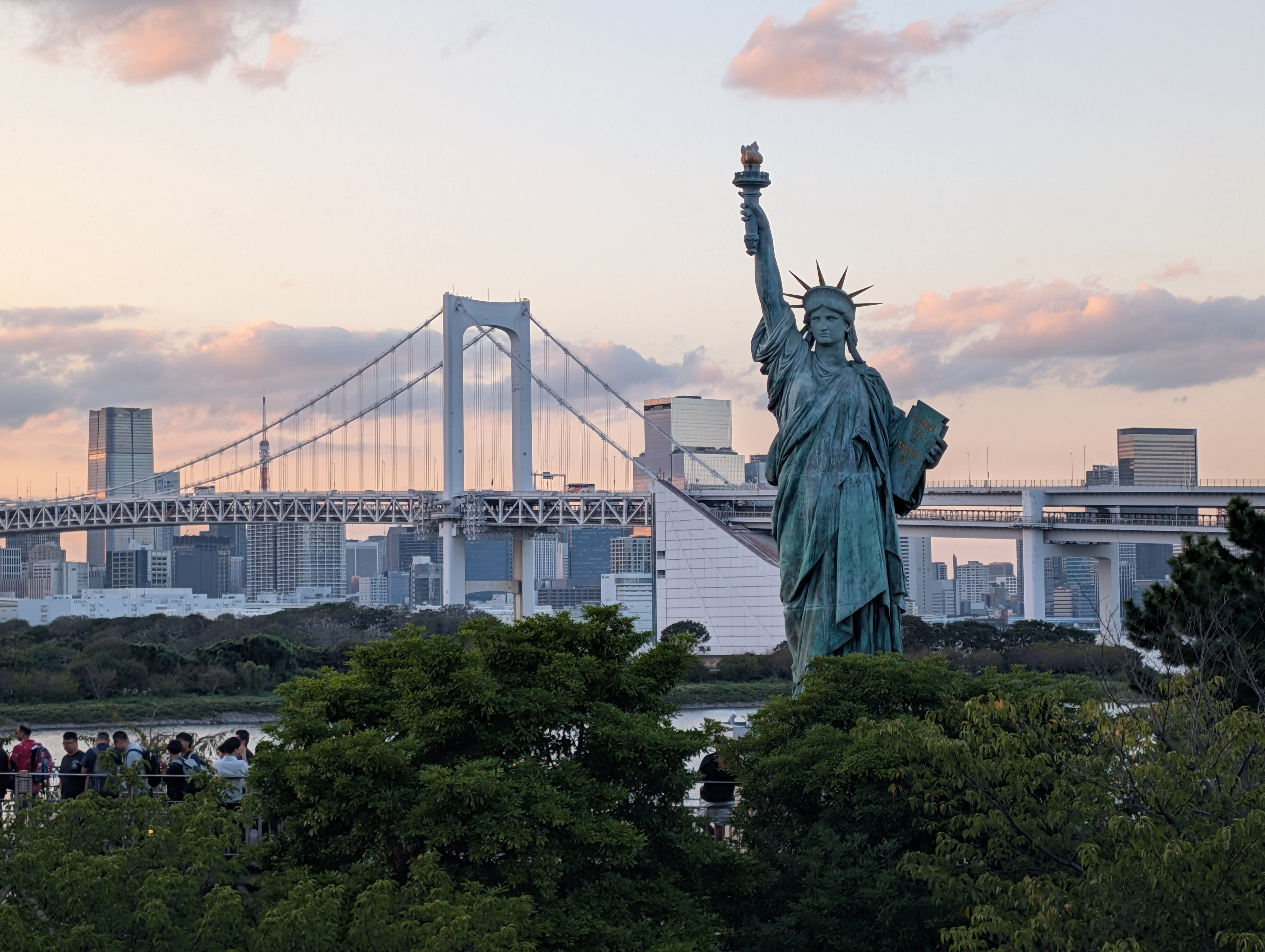 The Statue of Liberty replica at Odaiba at sunset — the Rainbow Bridge behind it, Tokyo's skyline in the golden distance