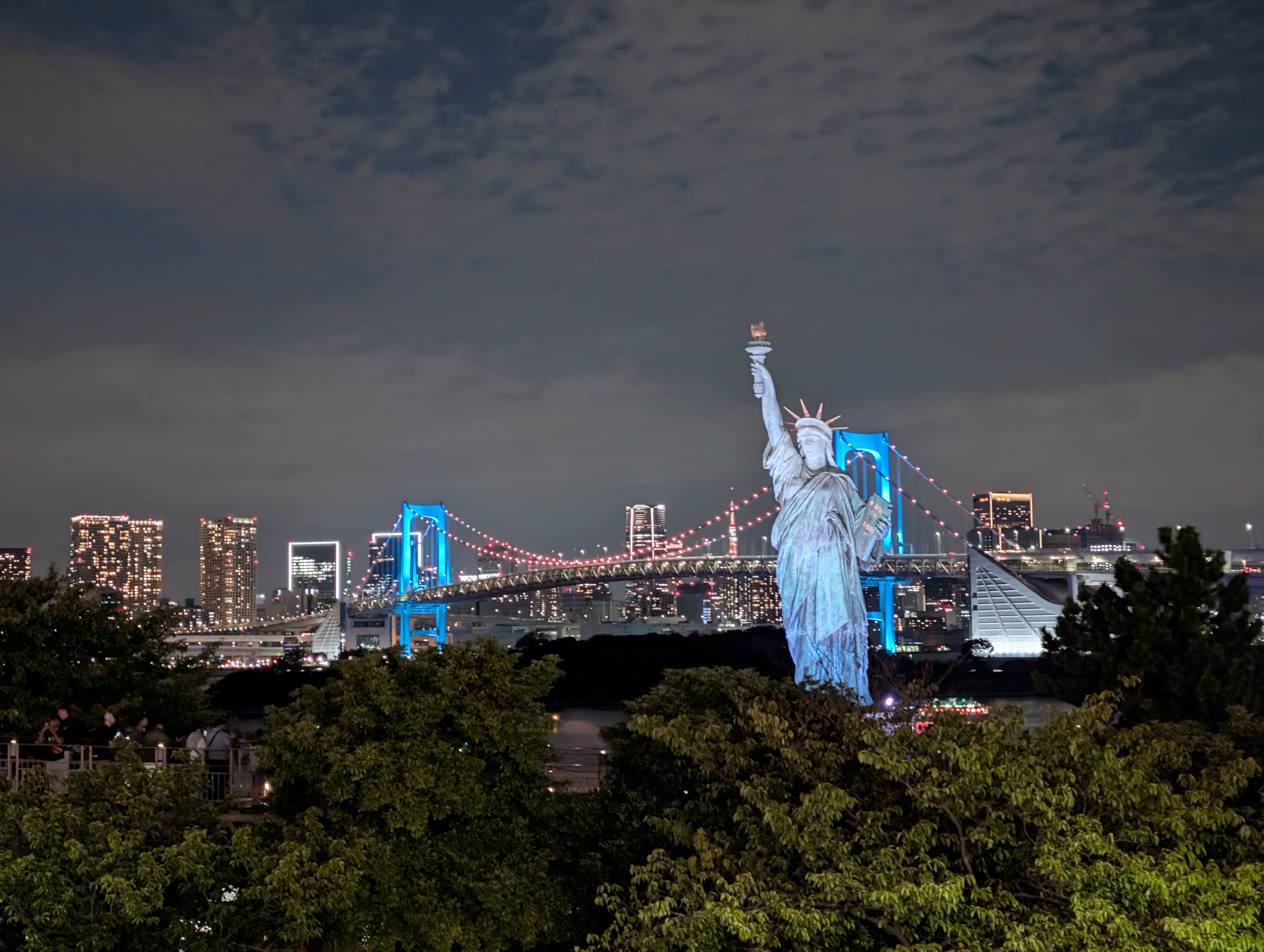 The Statue of Liberty and Rainbow Bridge at night — the bridge lit in blue, the city glowing, everything reflected in Tokyo Bay