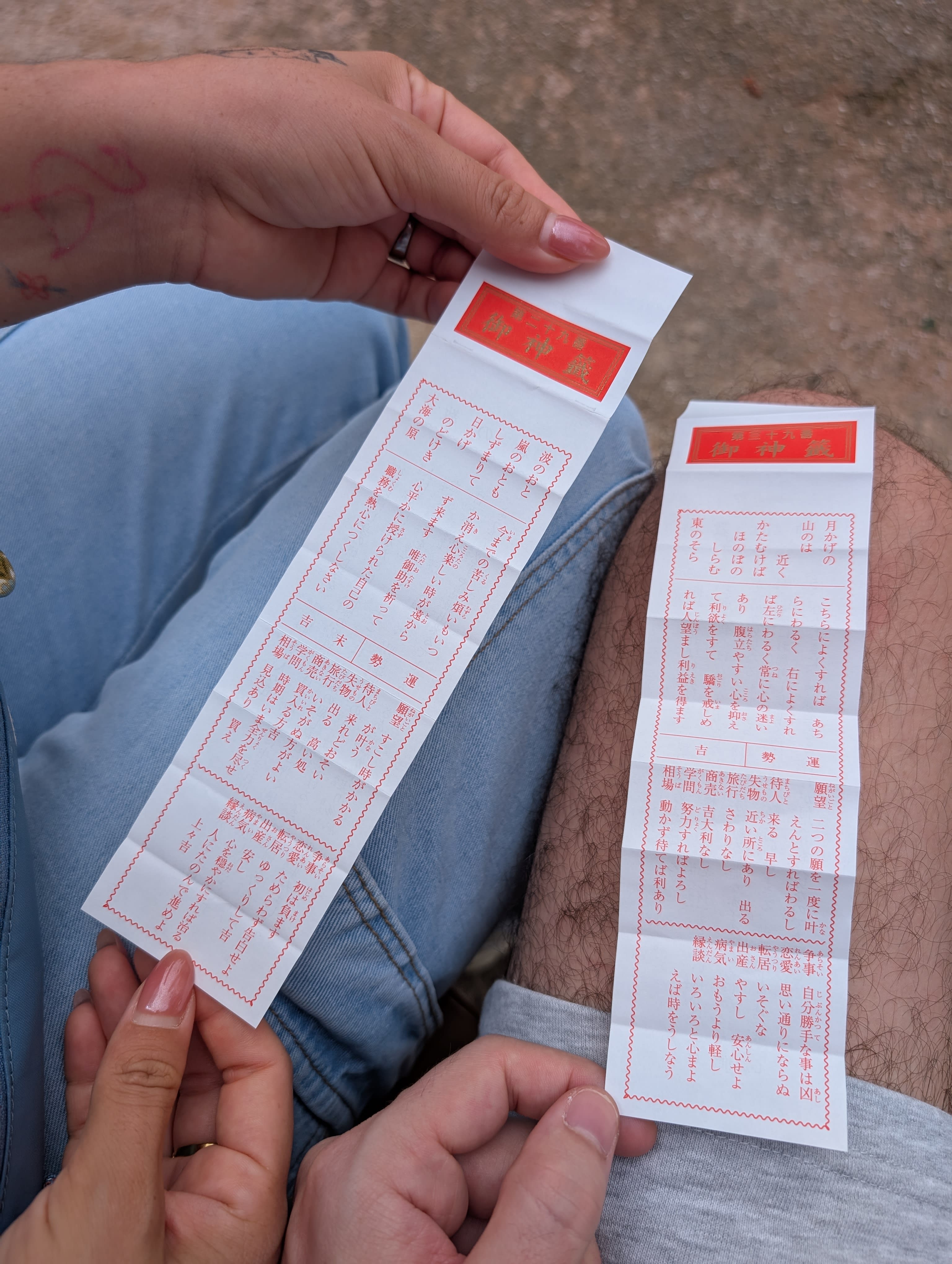 Two omikuji fortune papers held side by side — red and white text in Japanese kanji, resting on our laps at the temple