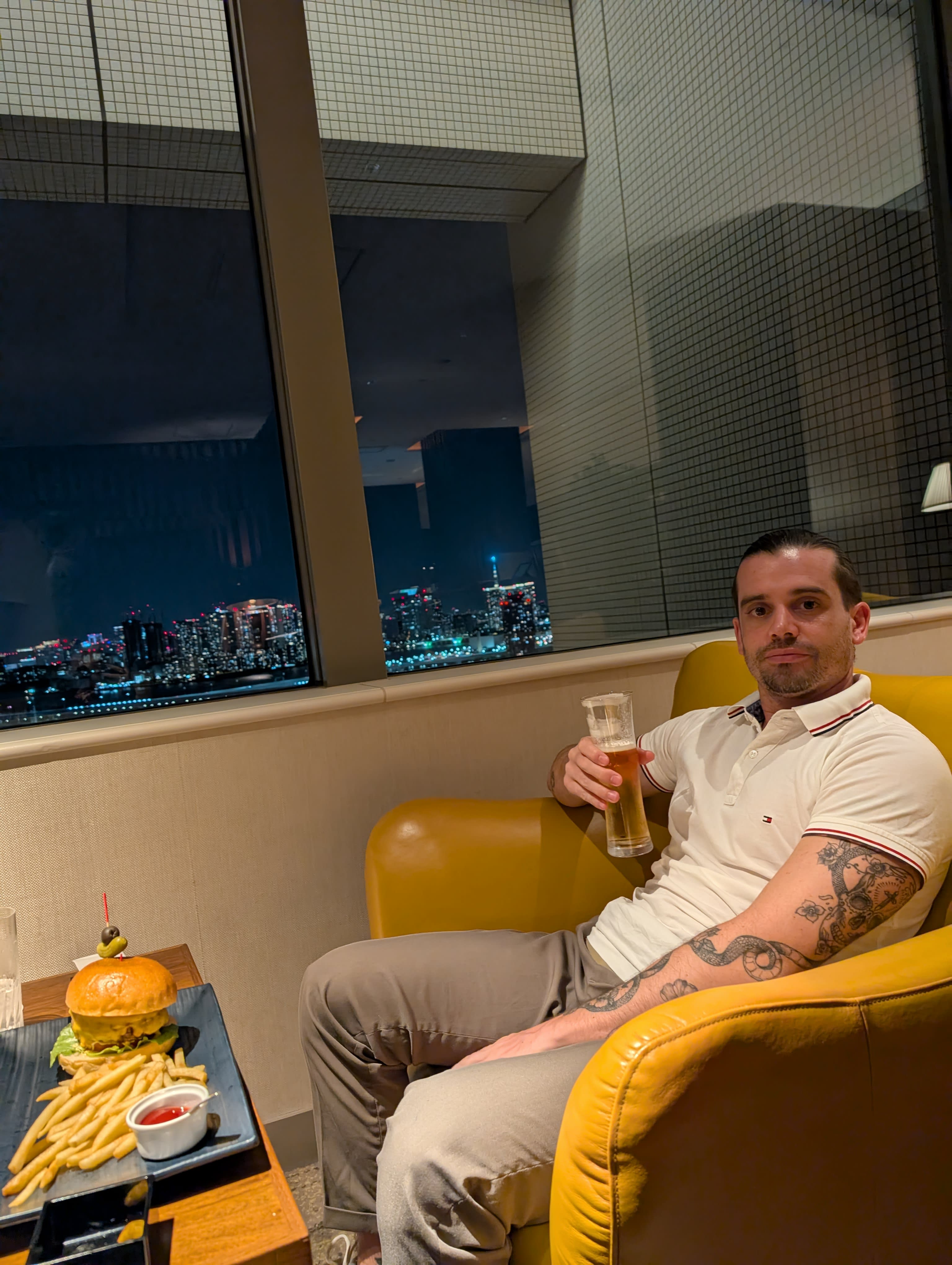 Pierre in the hotel lounge — yellow armchair, beer in hand, a burger and fries on the side table, Tokyo Bay's lights glowing through the window at night