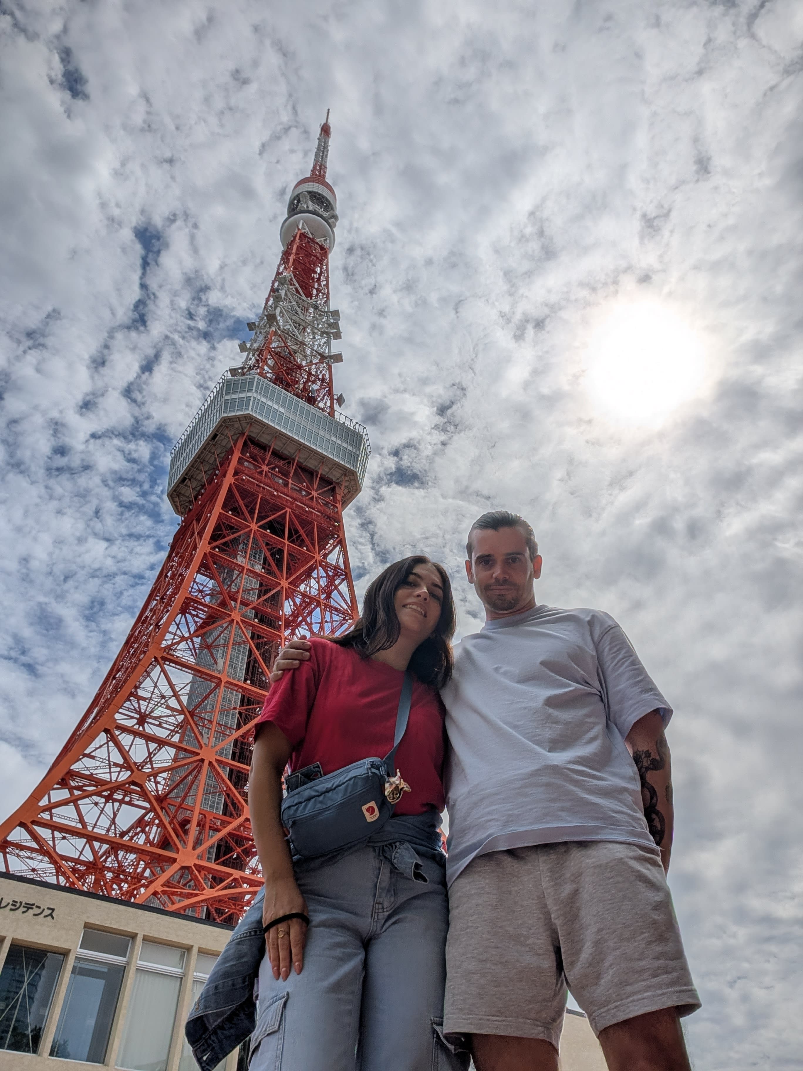 Pierre and Lia at the base of Tokyo Tower — looking up through the red steel lattice, clouds and sun behind, both smiling