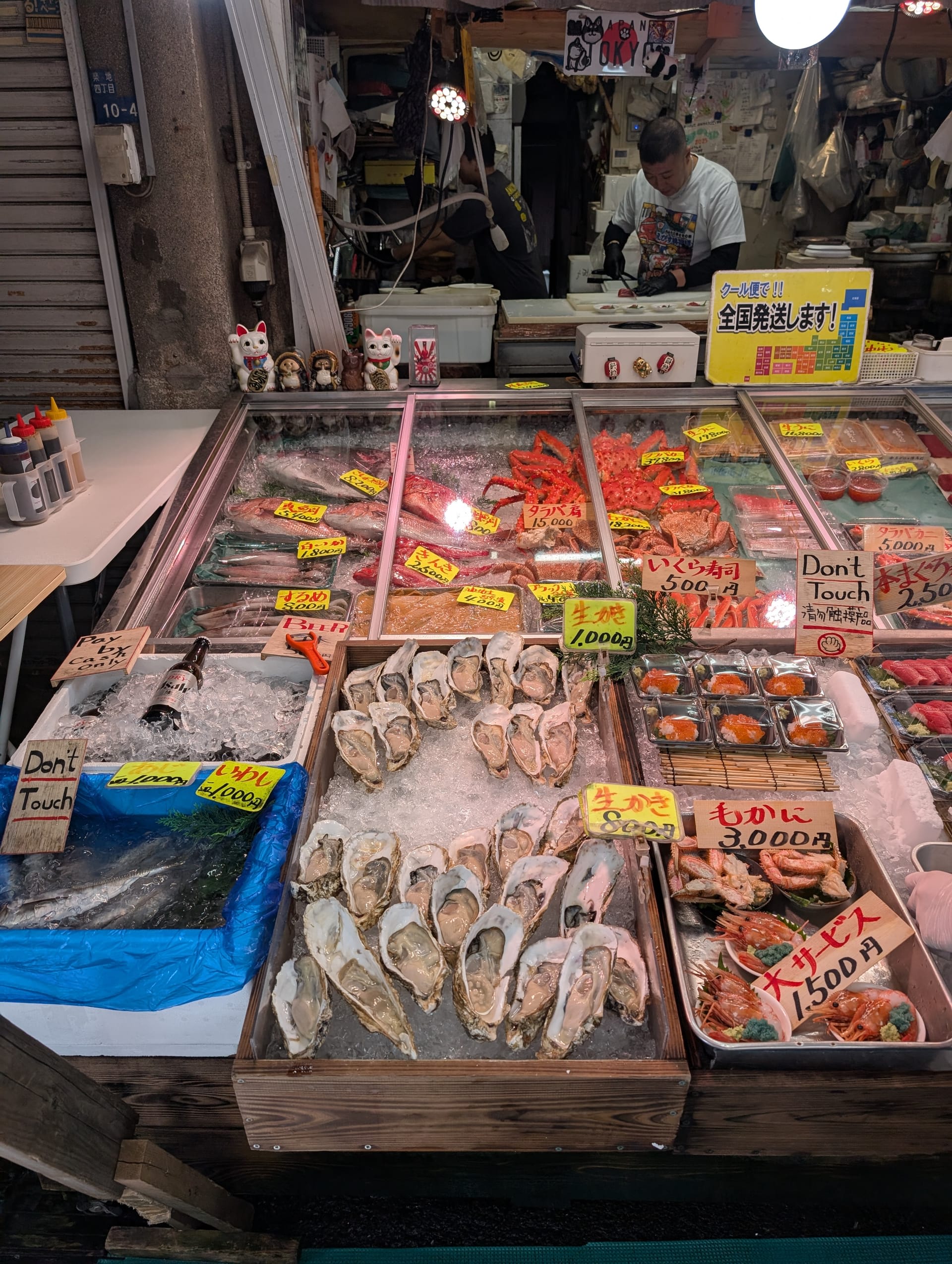 A Tsukiji stall displaying fresh oysters, crab legs, and seafood on ice — handwritten price tags, a vendor working in the background