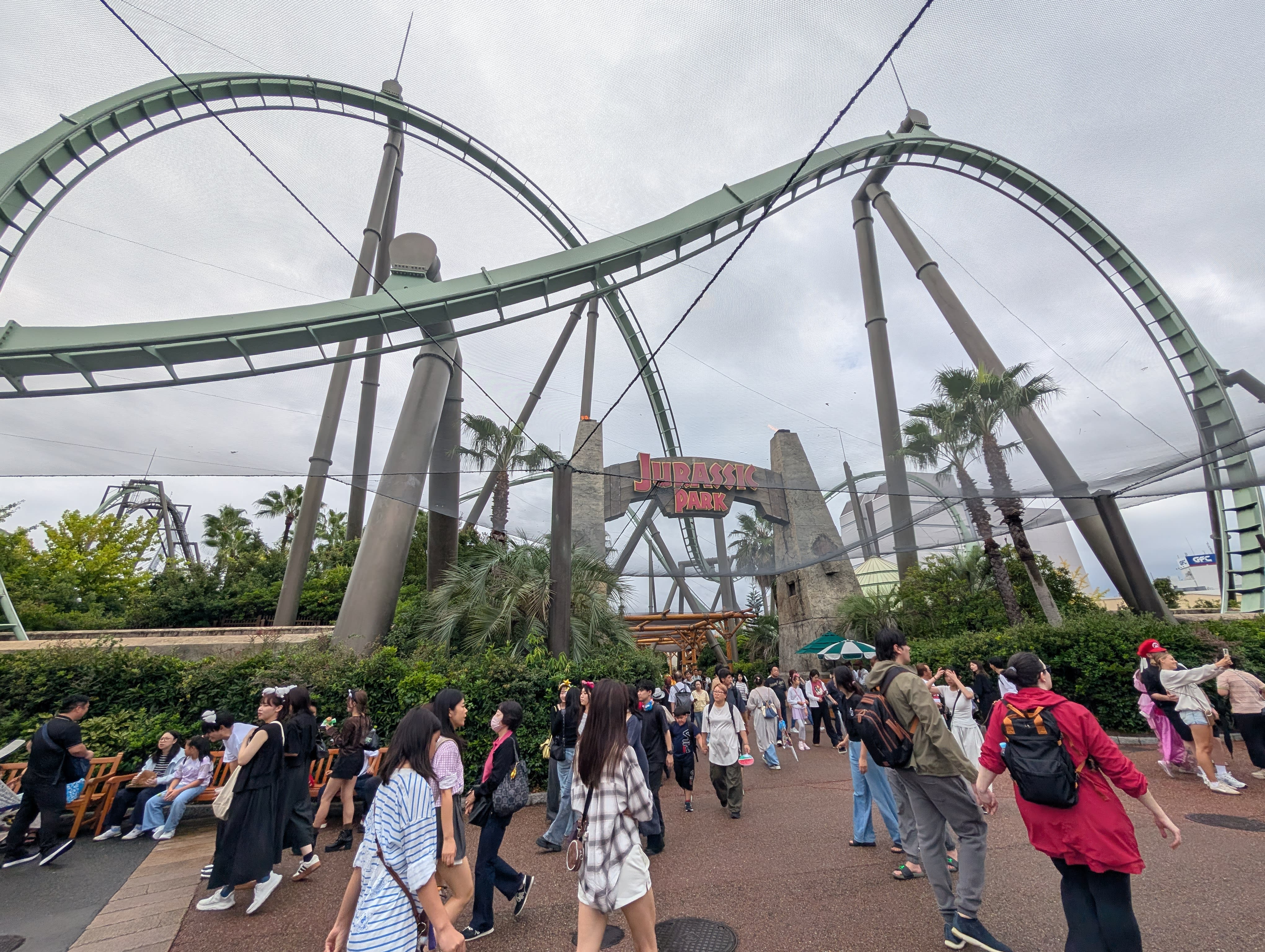 The Jurassic Park area — rollercoaster loops twisting overhead, the wooden gate, palm trees, crowds flowing through the walkway below