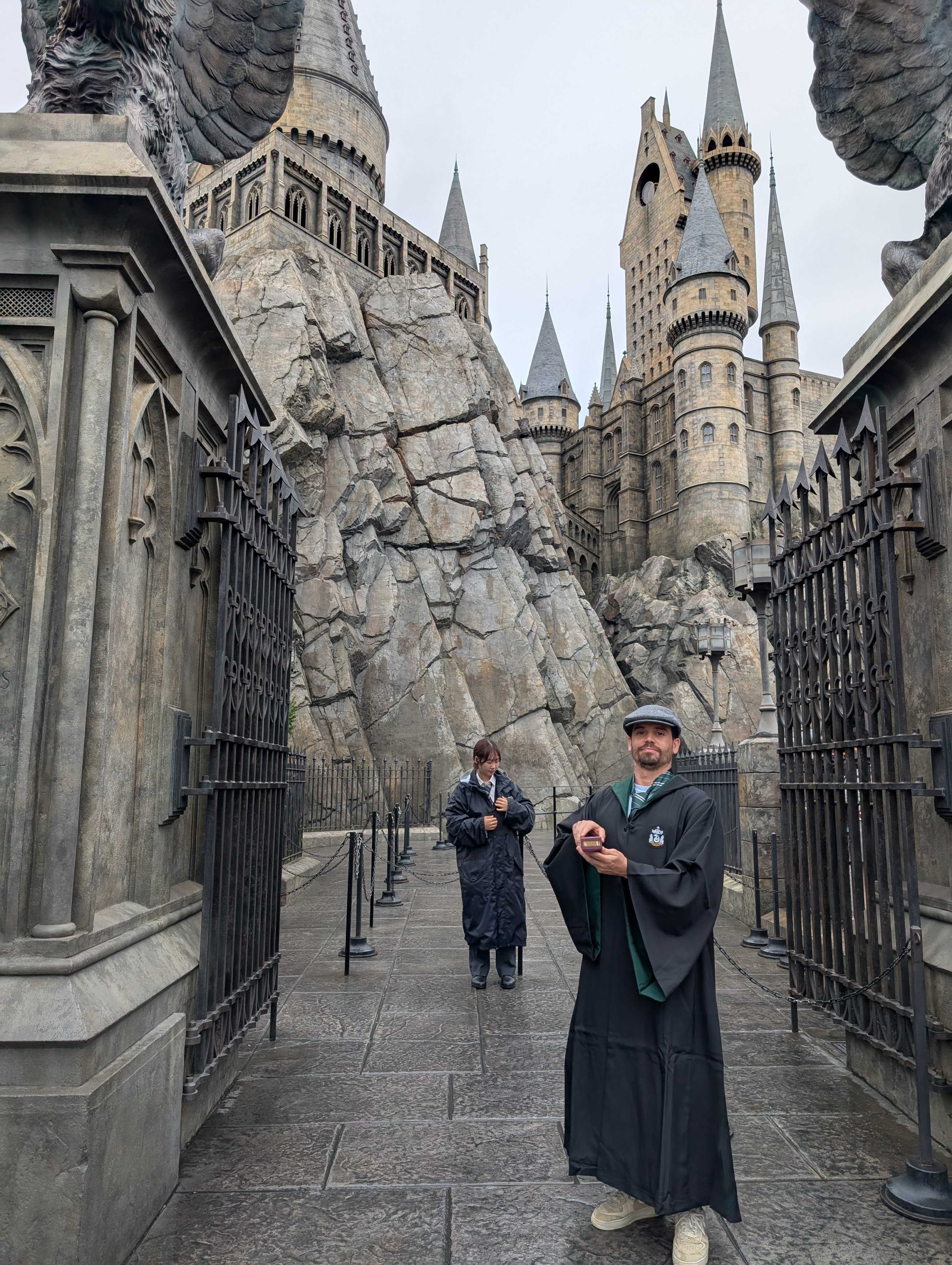 Pierre in full Slytherin robes on the bridge to Hogwarts Castle — the towers and turrets rising behind him, the grey stone, the sense that this is entirely too emotional for a grown man