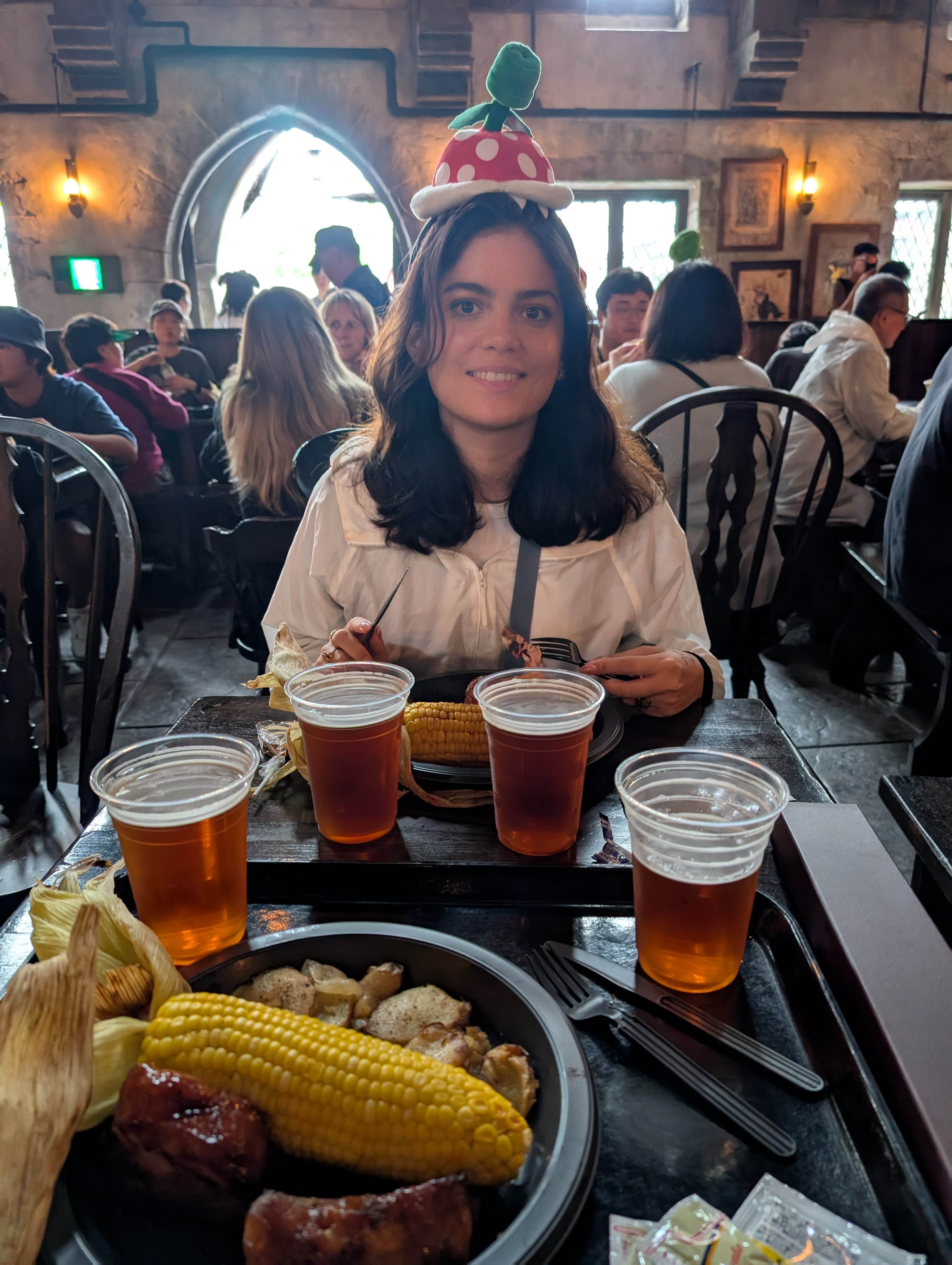 Lia at the Three Broomsticks — four butterbeers on the table, ribs and corn in front of her, the Piranha Plant hat still on, the medieval pub atmosphere glowing behind her