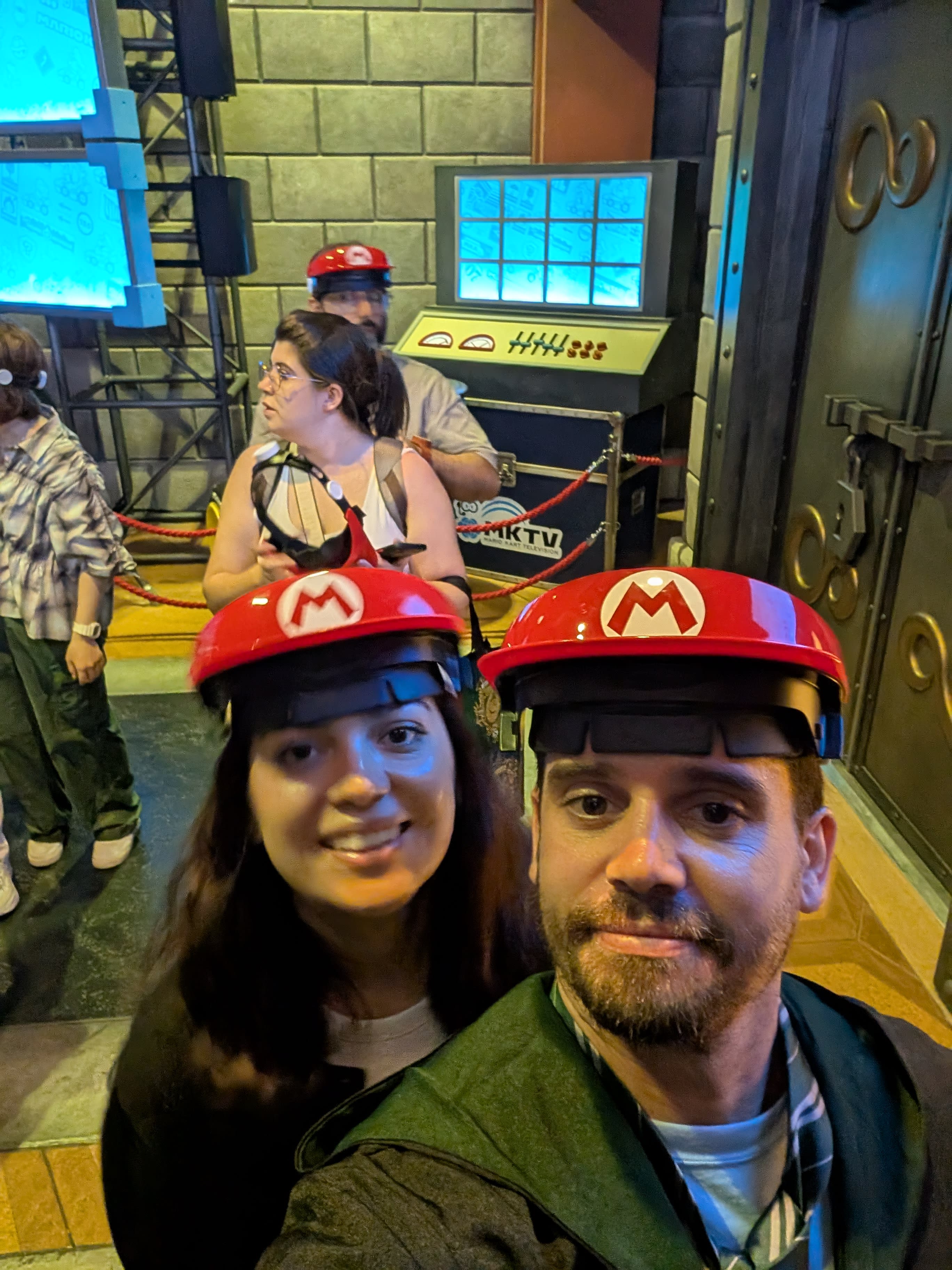 Pierre and Lia in Mario Kart helmets in the ride queue — the red caps with the M logo, the industrial set design behind them, two adults with zero dignity remaining