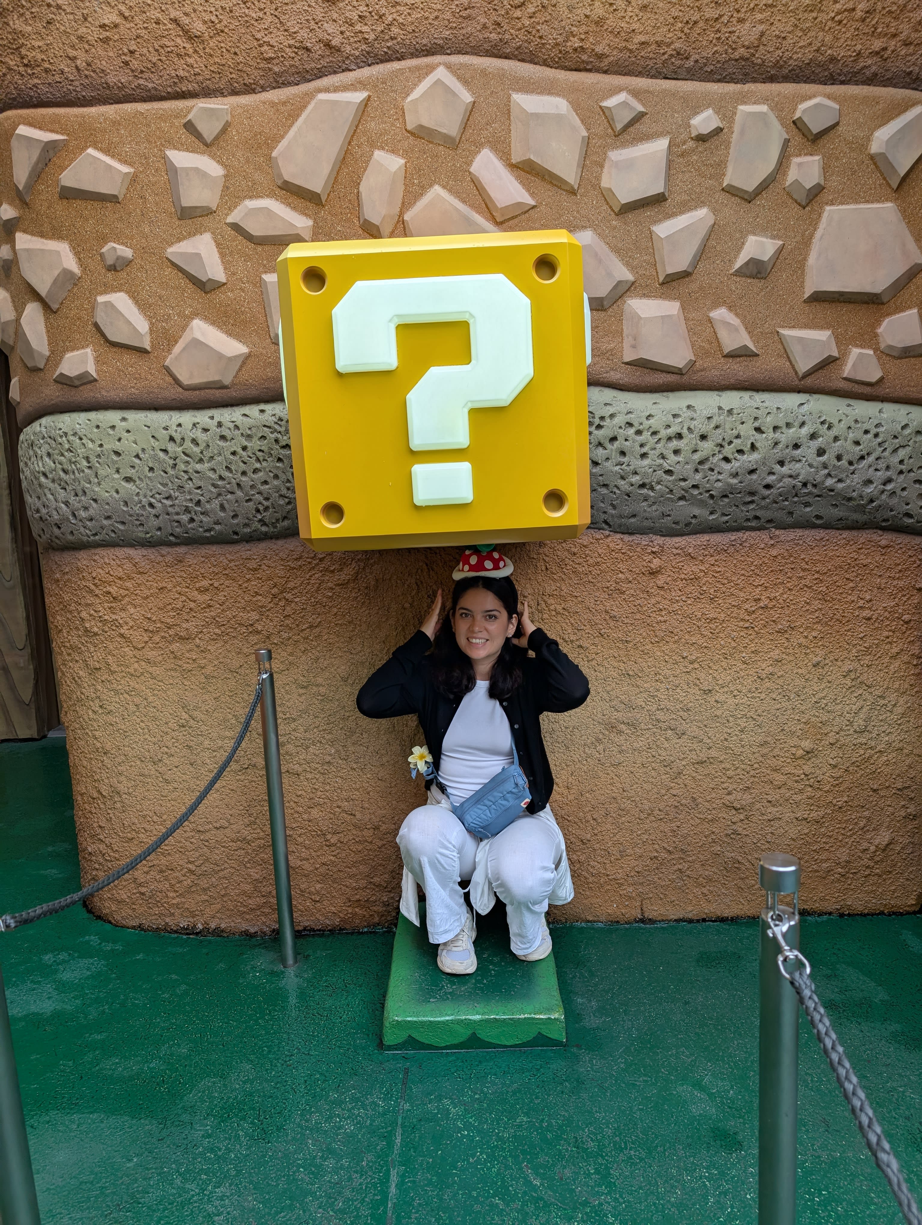 Lia crouching beneath a giant question block in Super Nintendo World — hitting it with her fist, the yellow block above her, the green mushroom floor beneath, pure joy
