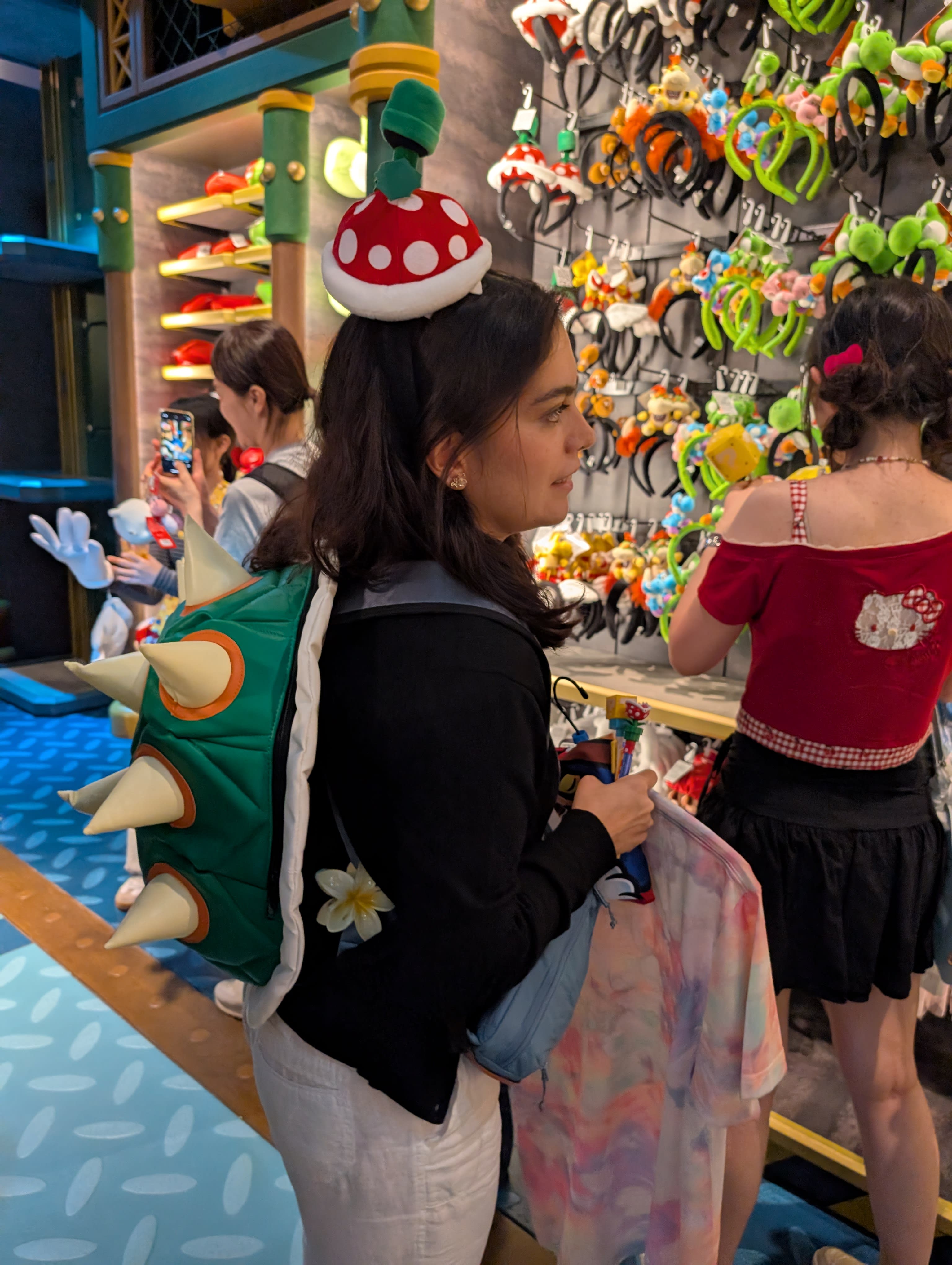Lia shopping in the Nintendo World store — Bowser shell backpack on, Piranha Plant hat on, examining character headbands on a wall display with absolute seriousness