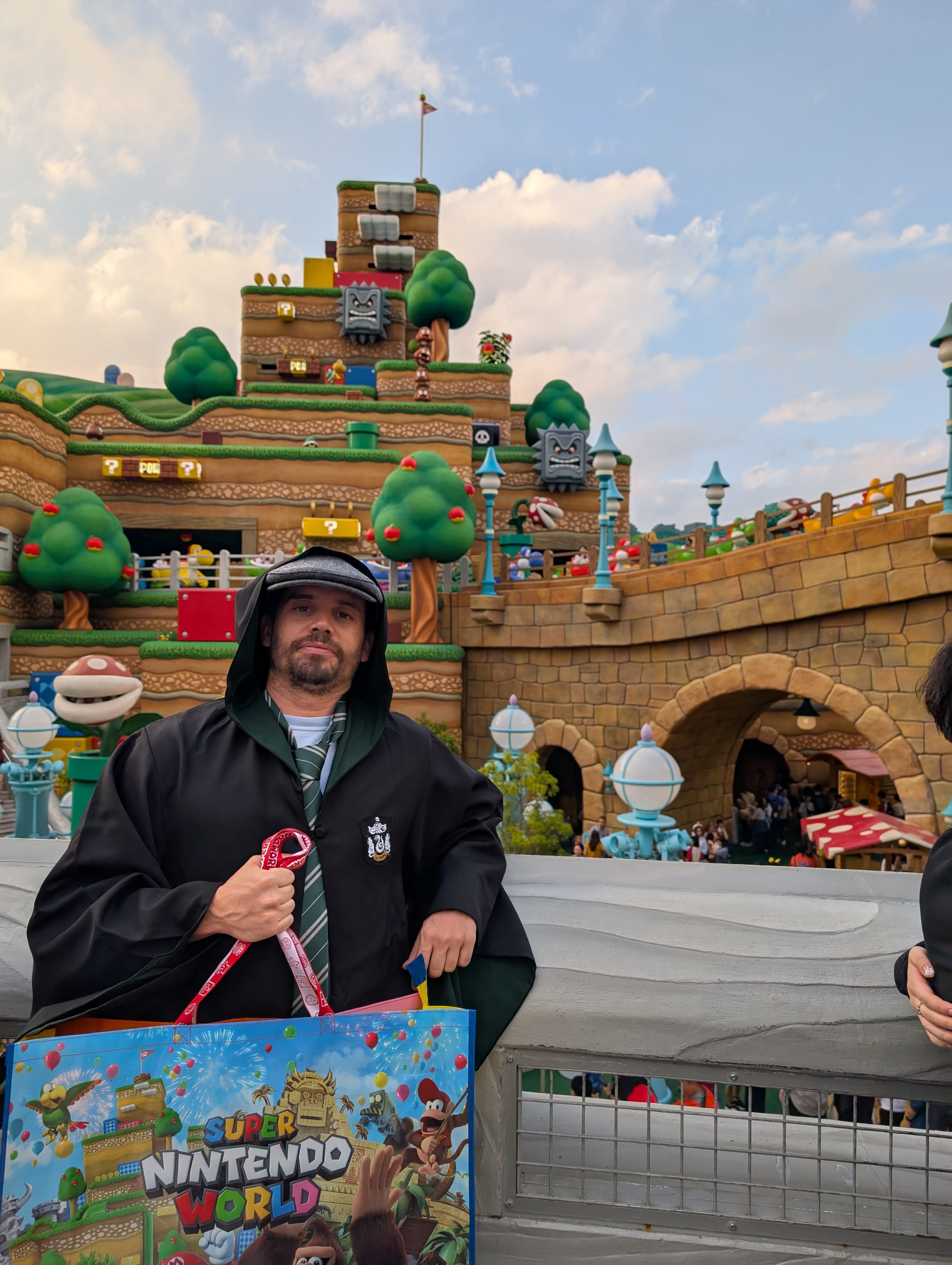 Pierre at the Nintendo World overlook in Slytherin robes — the full Mushroom Kingdom spread behind him, Bowser's Castle on the hill, a Nintendo shopping bag in hand, franchise loyalty divided