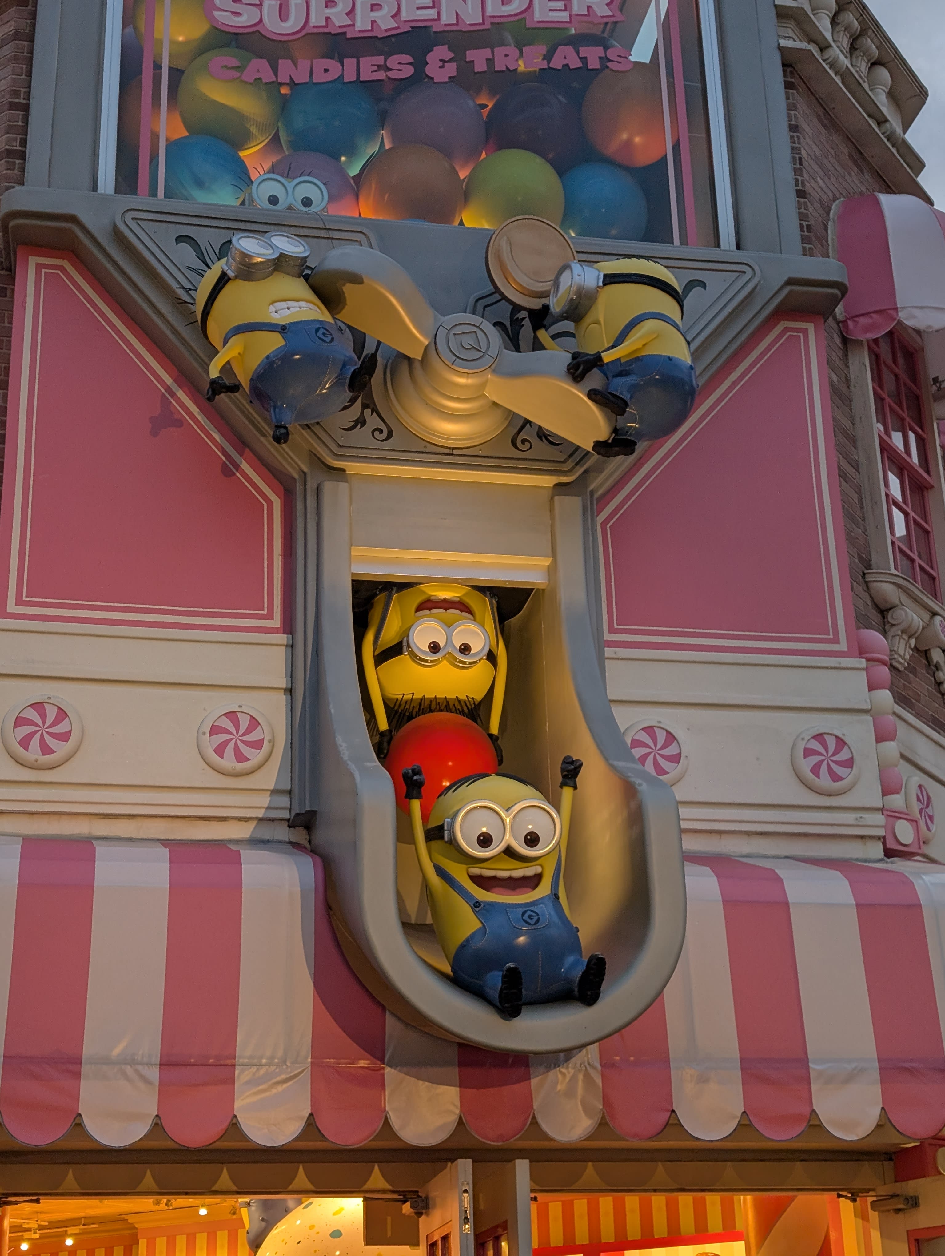The Surrender Candies & Treats shop — Minions tumbling out of a window and sliding down a chute, the pink facade, balloons above, candy-shop awnings below