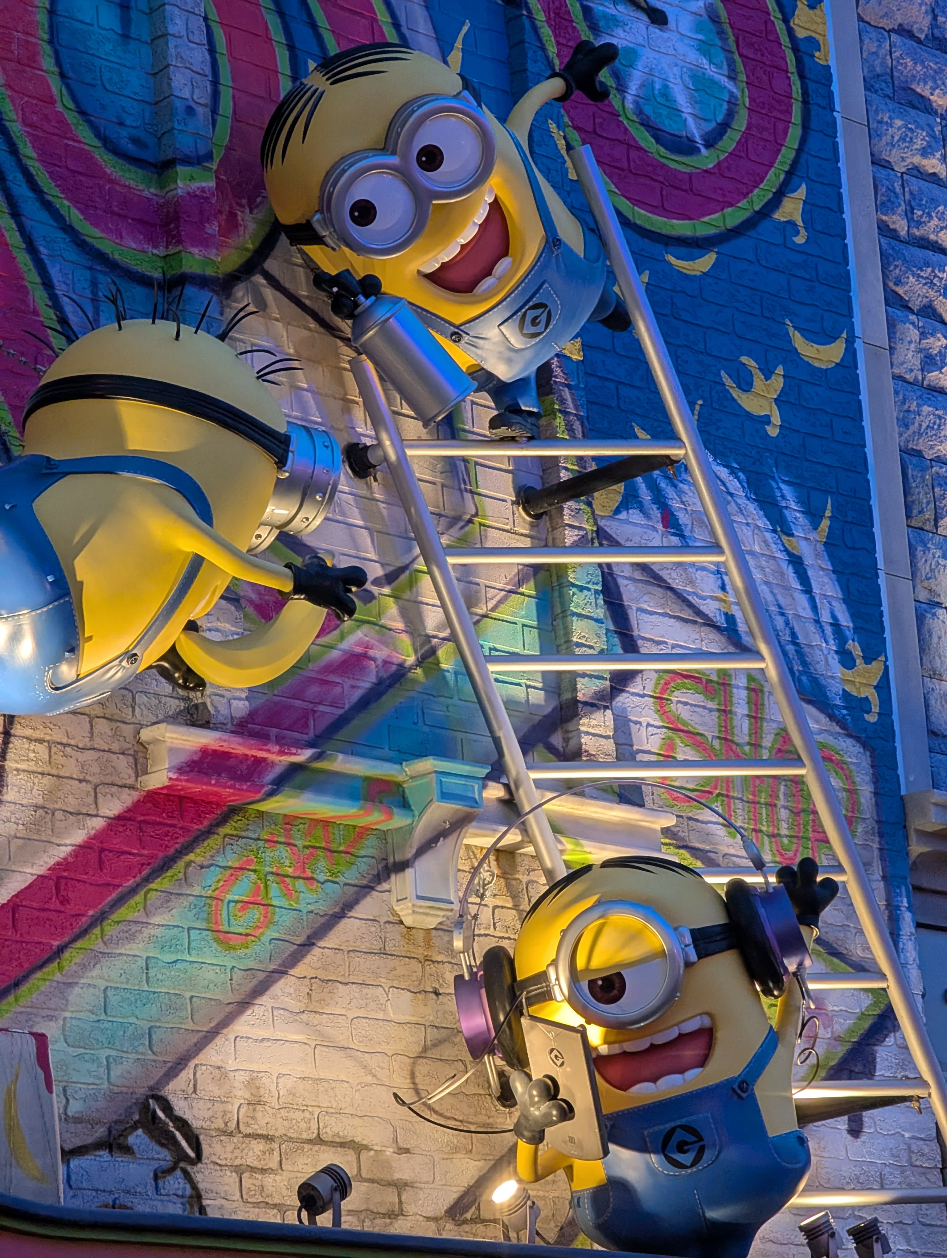 Minions climbing a ladder on a graffiti wall — the colours popping against the dusk sky, the yellow and blue and purple of their overalls vivid against the street art