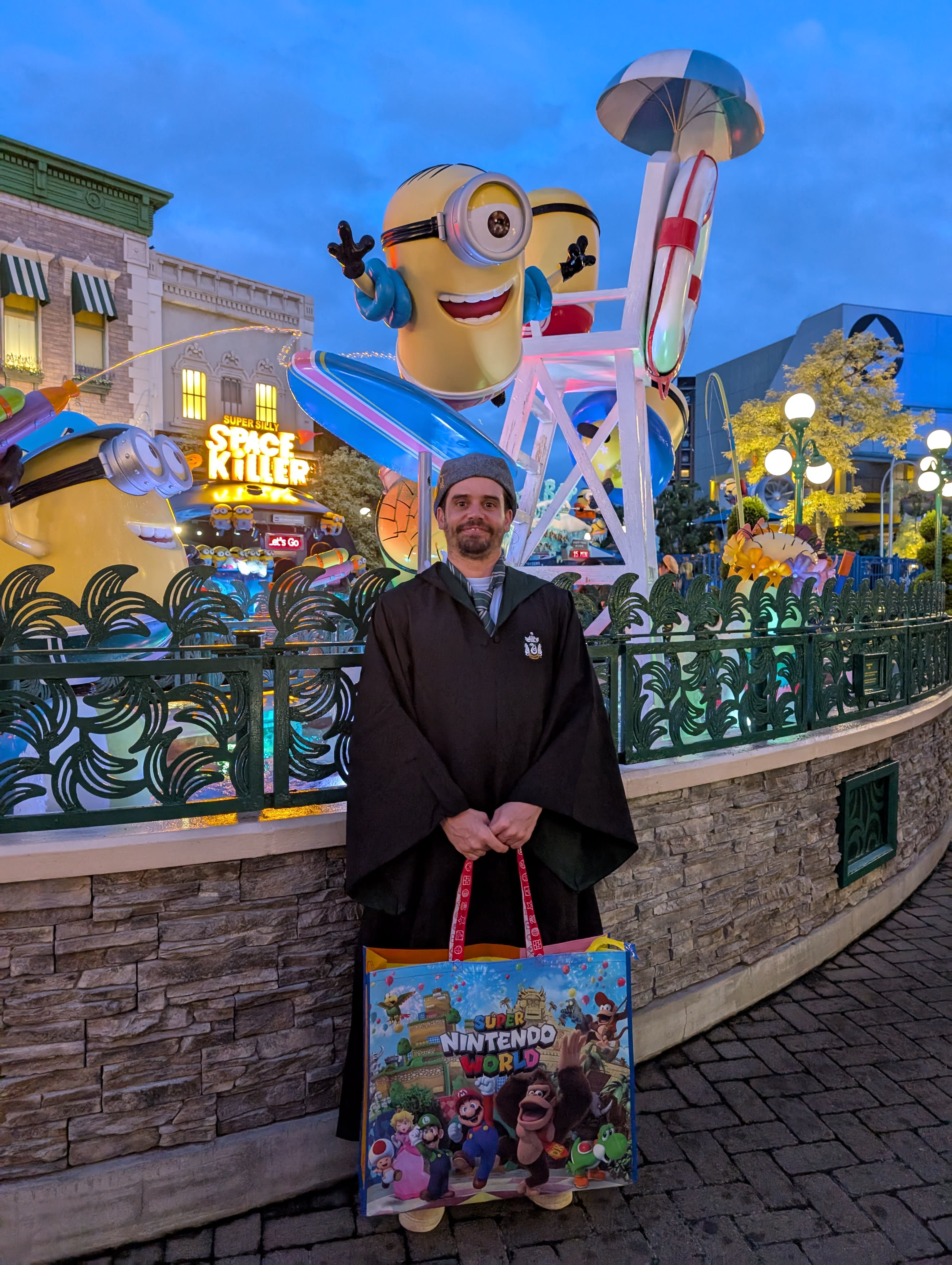 Pierre in Slytherin robes at Minion Park at blue hour — a giant Minion waving behind him, the Space Killer ride glowing, the Nintendo bag completing the cross-franchise ensemble