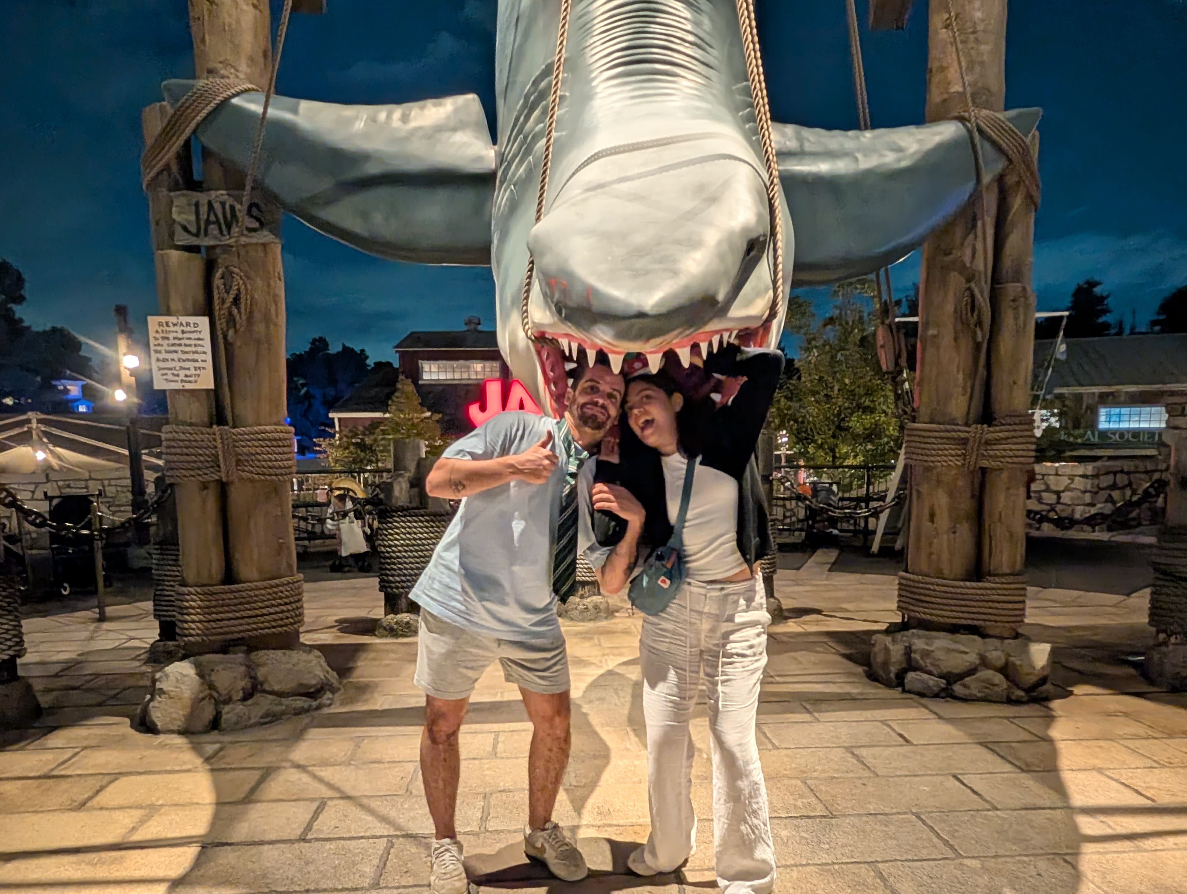 Pierre and Lia beneath the JAWS shark at night — Lia mid-scream, Pierre giving a thumbs up, twelve hours of theme park joy behind them