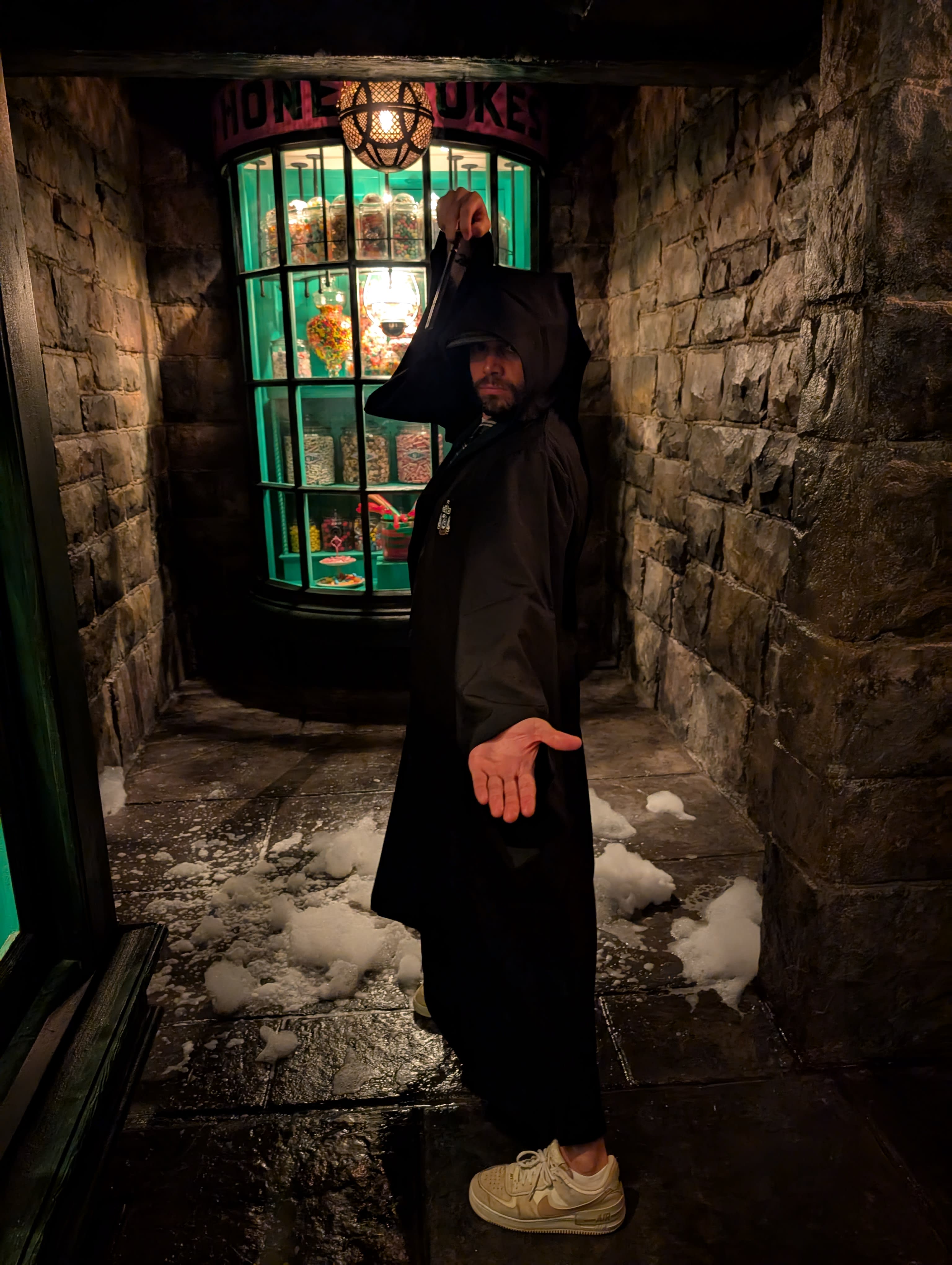 Pierre in full Slytherin robes in a snowy Hogsmeade alley at night — the Honeydukes shop glowing green behind him, fake snow on the cobblestones, full wizard commitment