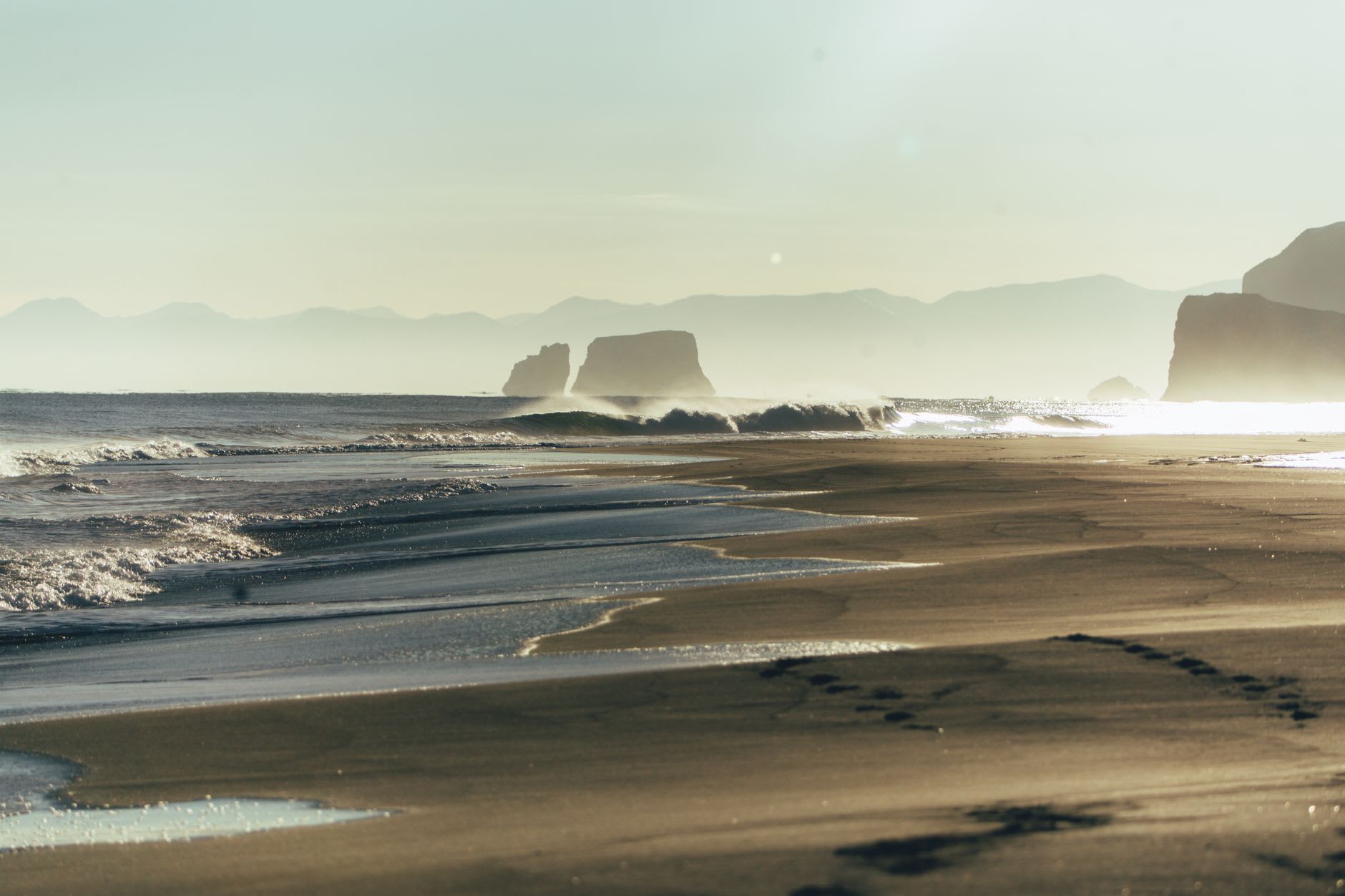 A lone set of footprints crossing the black sand of Khalaktyrsky Beach toward the breaking surf, the dunes and low coastal vegetation receding into mist