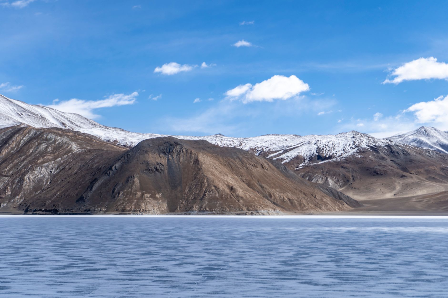Dawn light on Pangong Tso, the water golden-orange near the eastern end, prayer flags fluttering on a stone cairn in the foreground