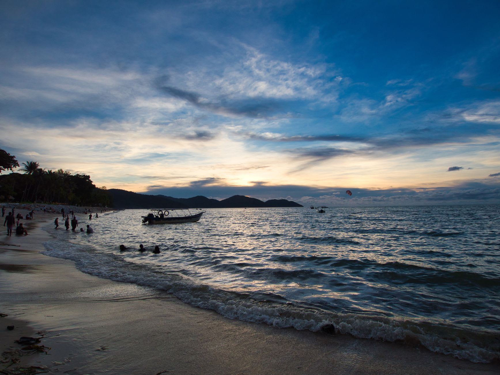 Swimmers in calm shallow turquoise water at Pantai Cenang at golden hour, palm trees silhouetted against the sunset sky, a long white beach stretching to the horizon