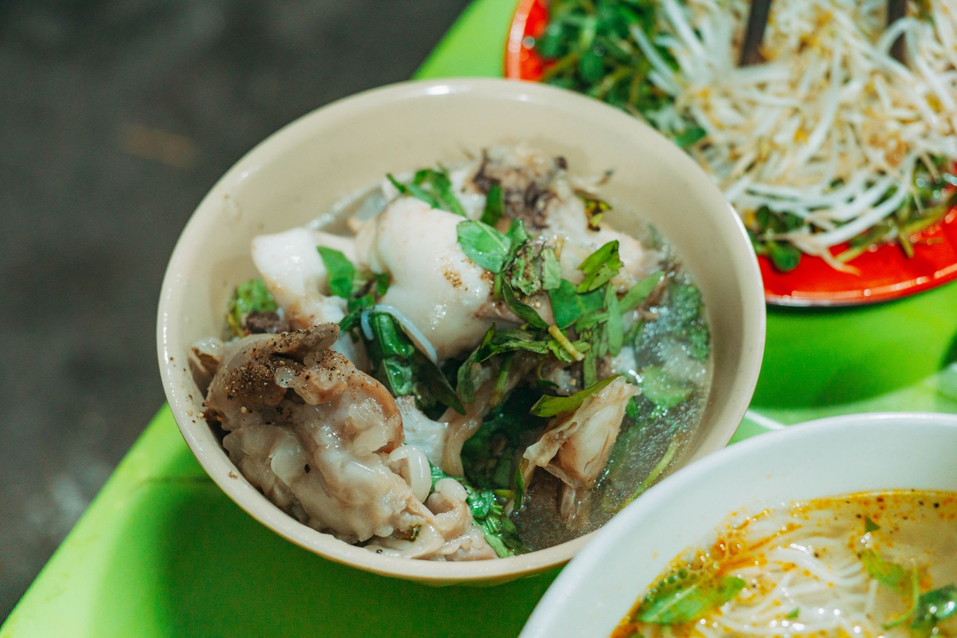 A bowl of khao piak sen at a street-side table on the Mekong waterfront, herbs and chillies on the side, the river visible behind